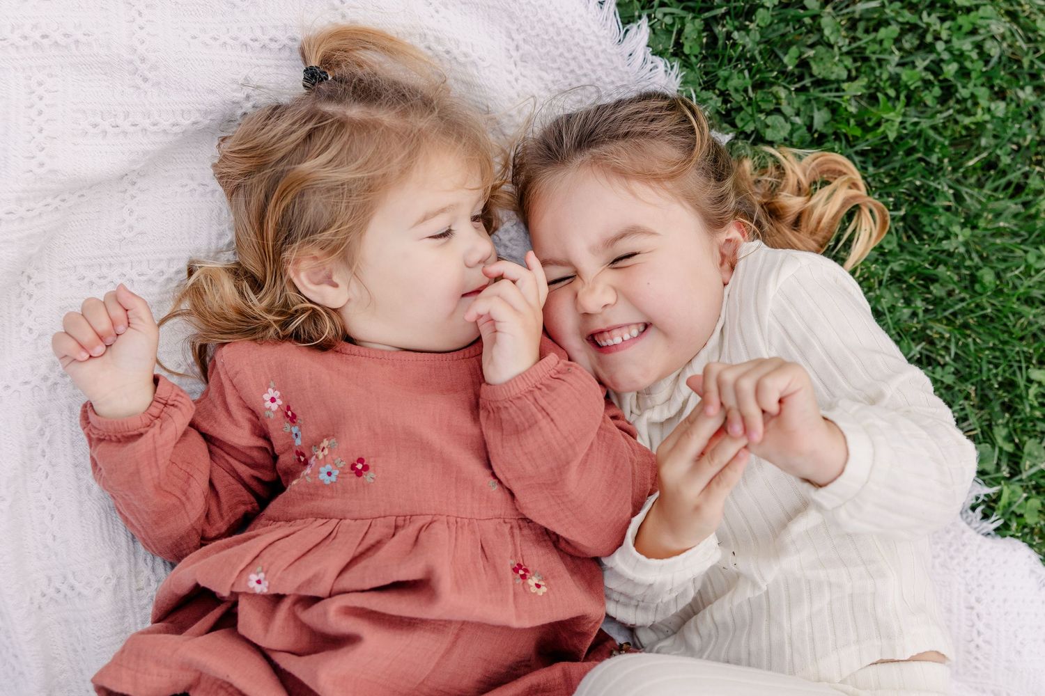 Two young girls in pink and white dresses laughing together while lying on a blanket outdoors.