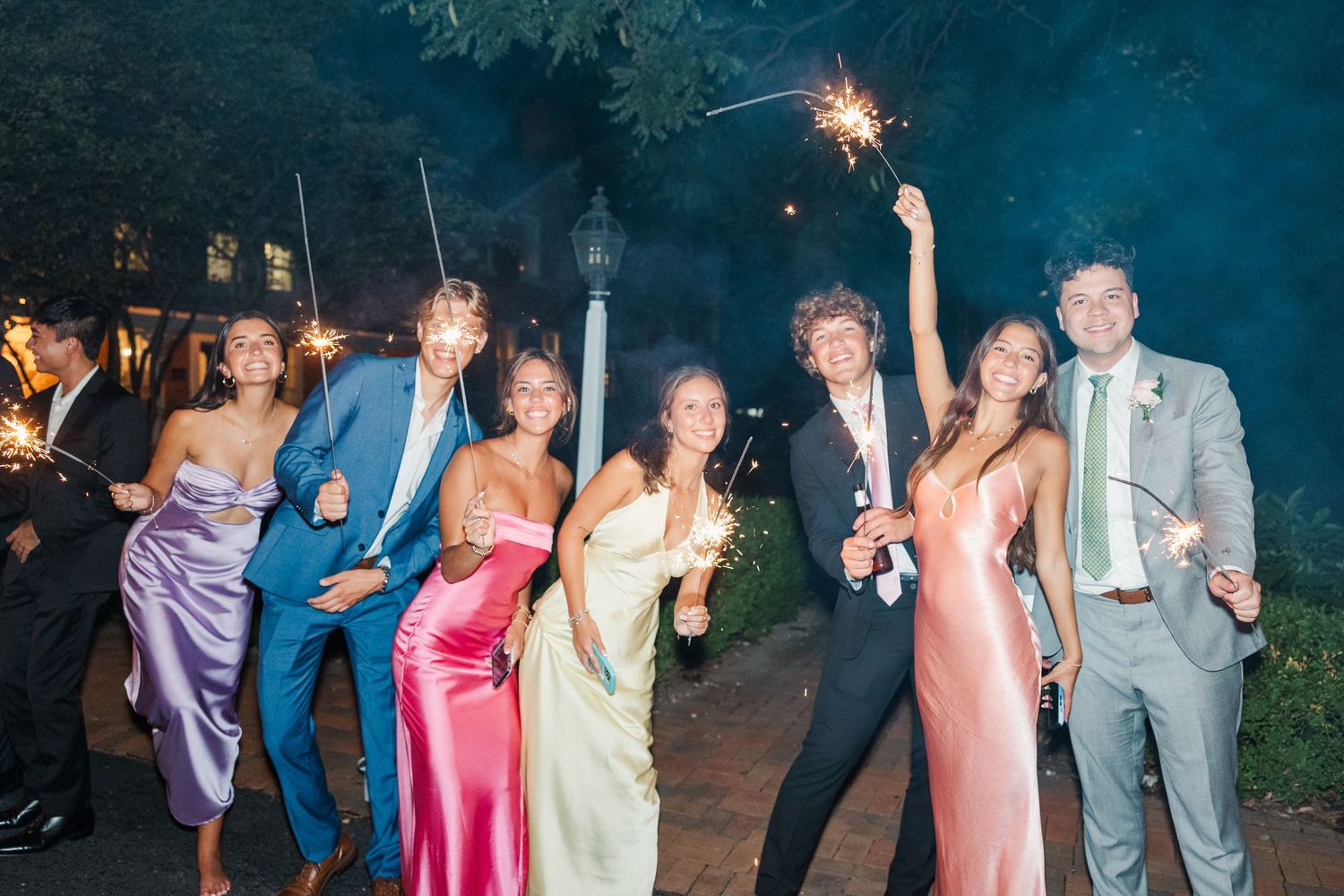 Wedding party members hold sparklers while posing outside at night in formal attire.
