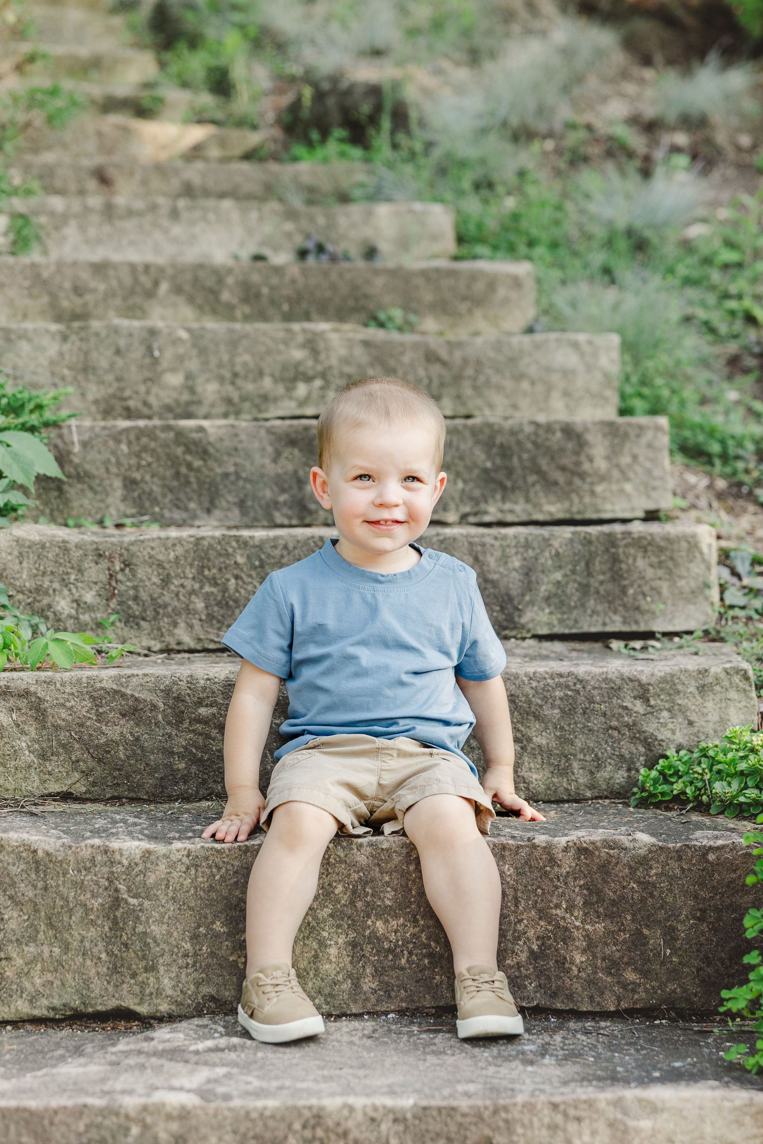 A small child in a blue t-shirt and beige shorts sits smiling on outdoor stone steps.