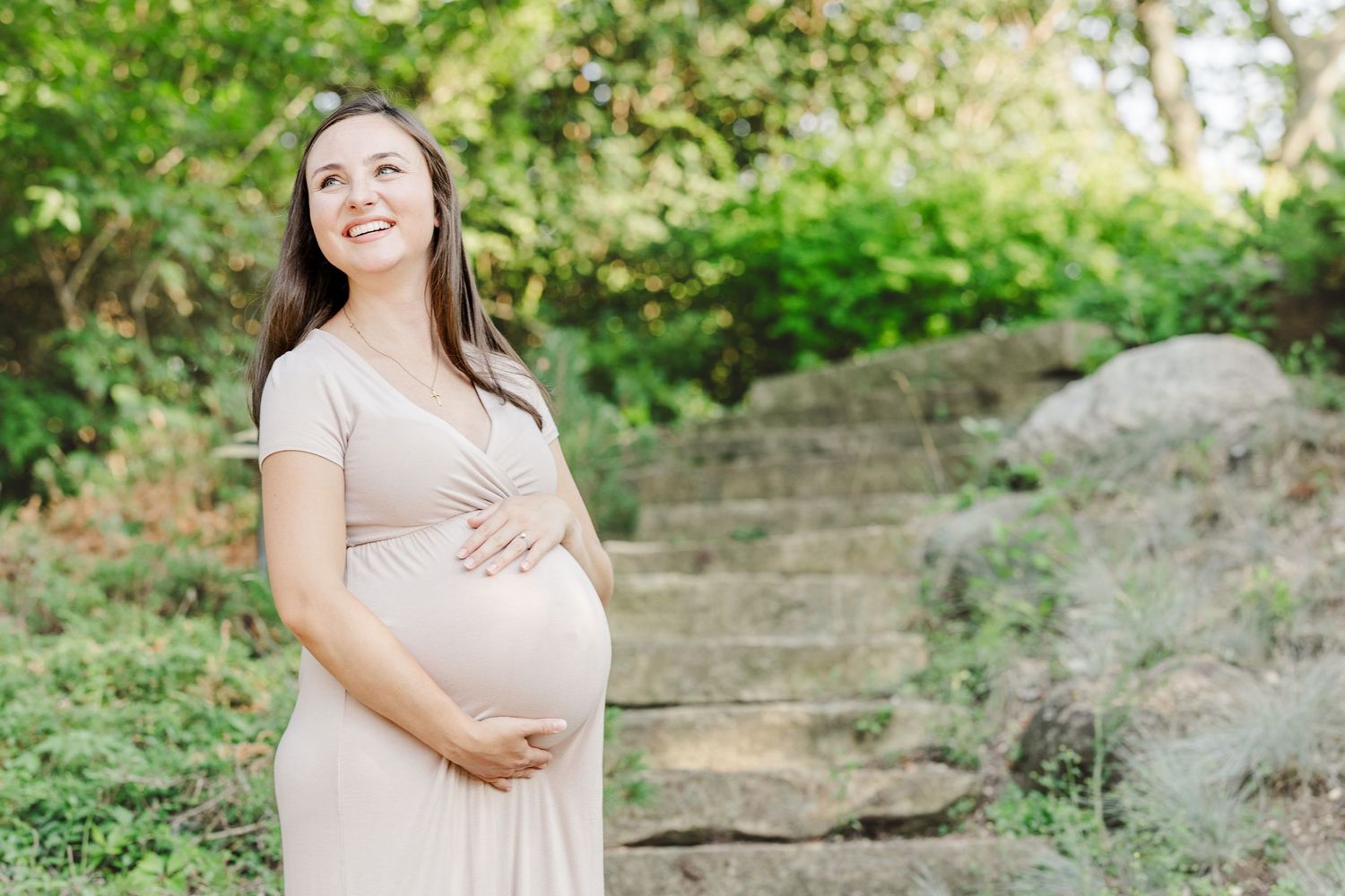 A pregnant woman in a flowing beige dress stands on outdoor stone steps surrounded by lush greenery.