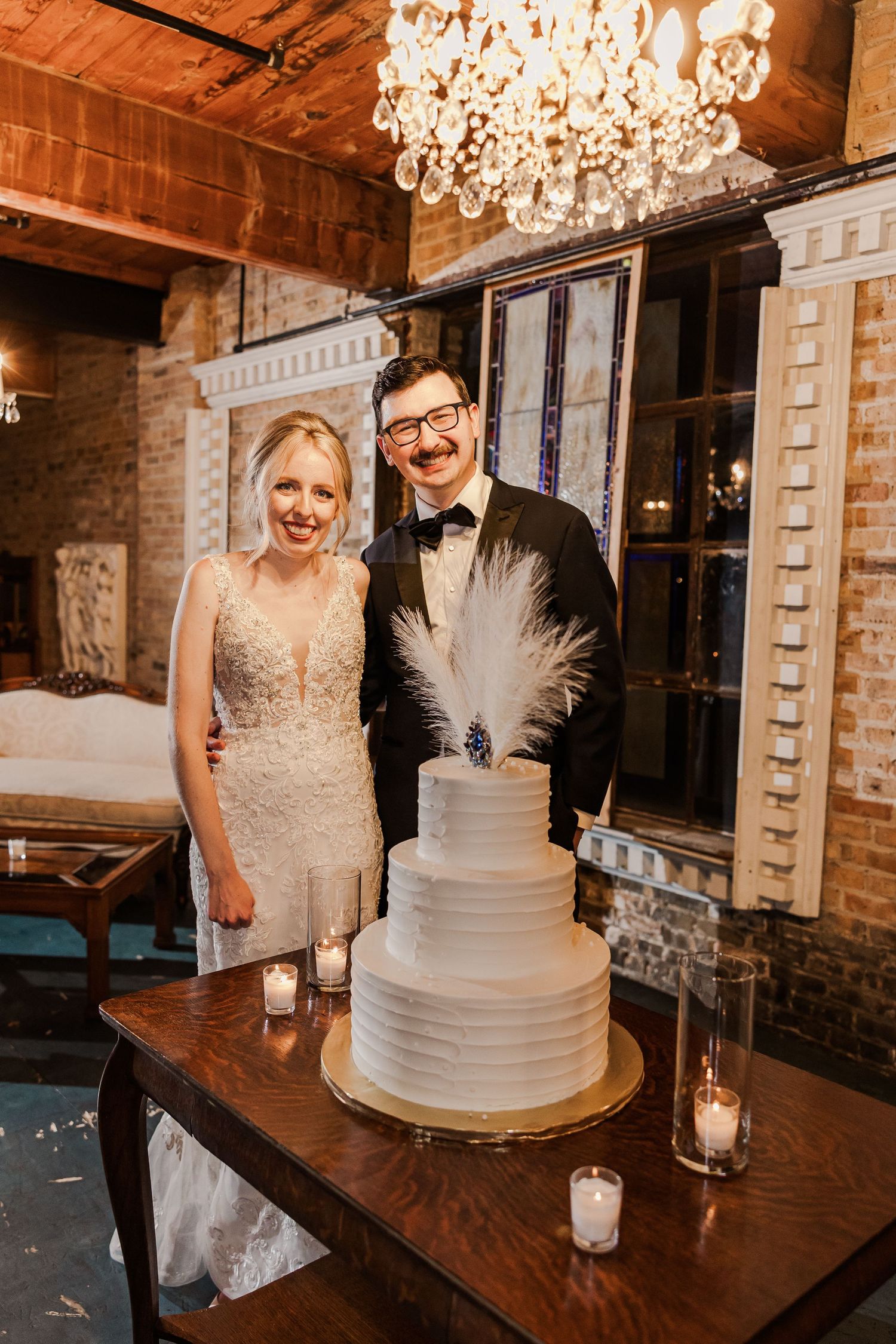 Newlyweds stand beside their elegant white wedding cake with feather topper under crystal chandelier.