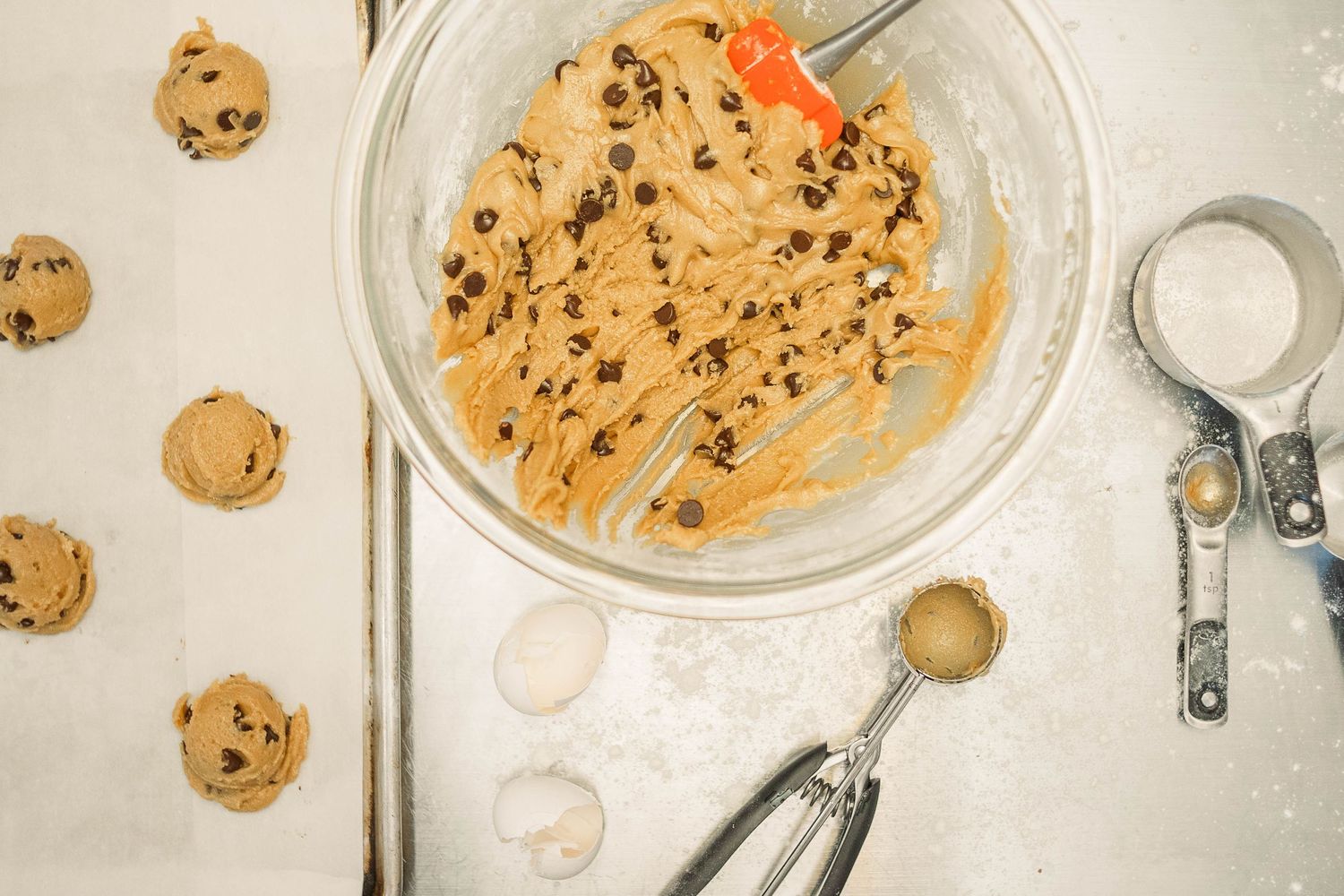 Cookie dough mixing bowl with chocolate chips and measuring spoons on white surface.