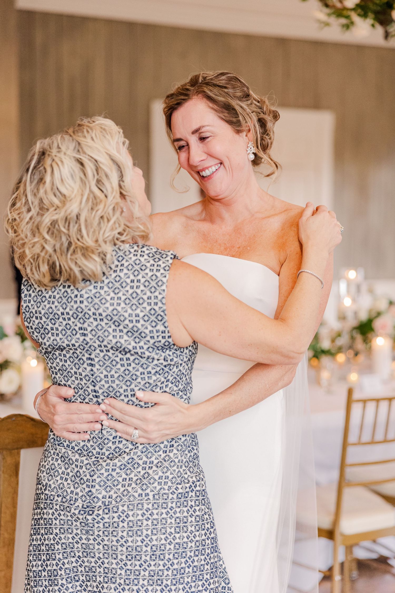 Two people share a tender embrace in a sunlit room with wedding reception decor.