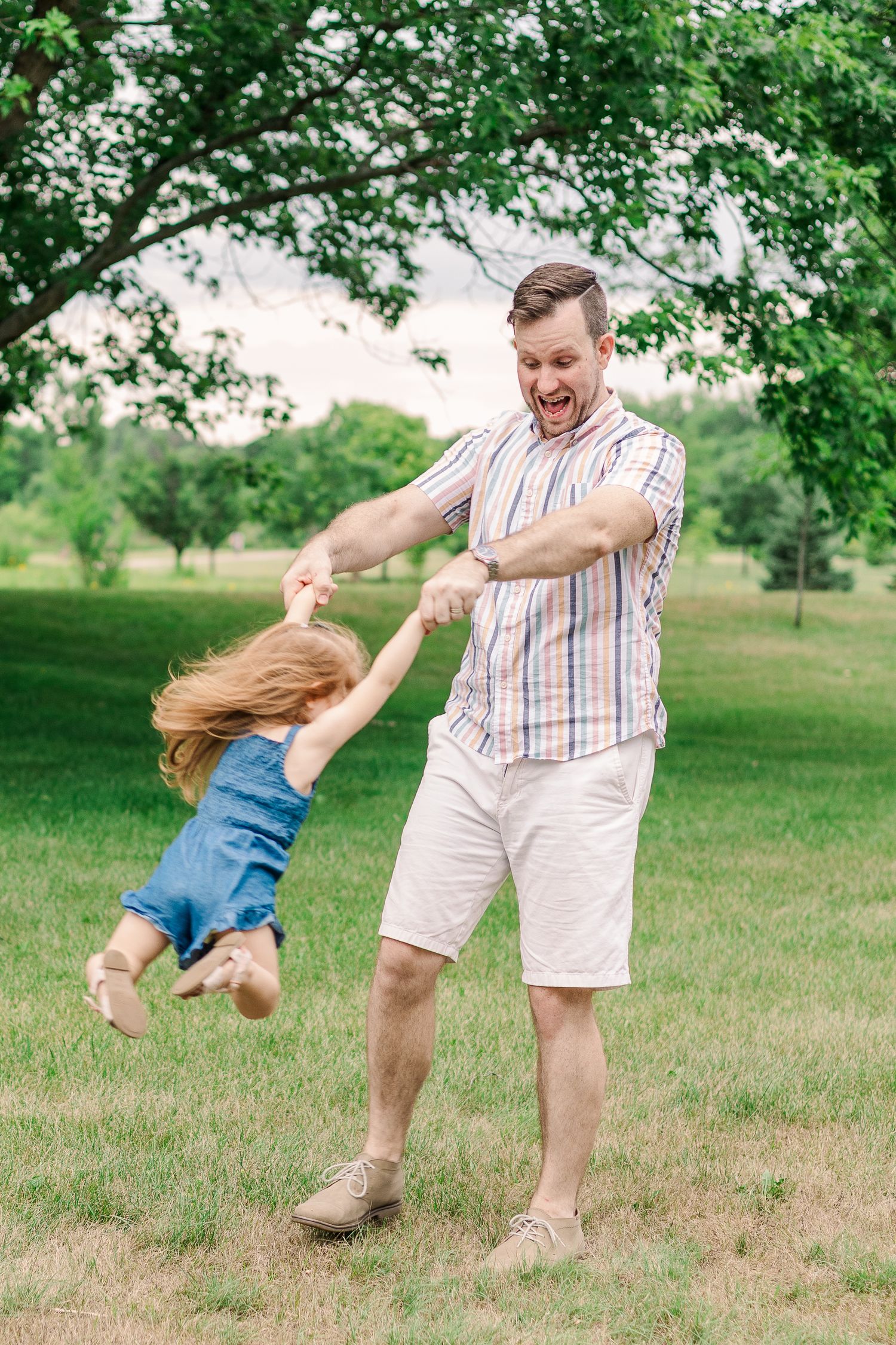 Playful outdoor scene showing someone spinning a child in a grassy field under trees.