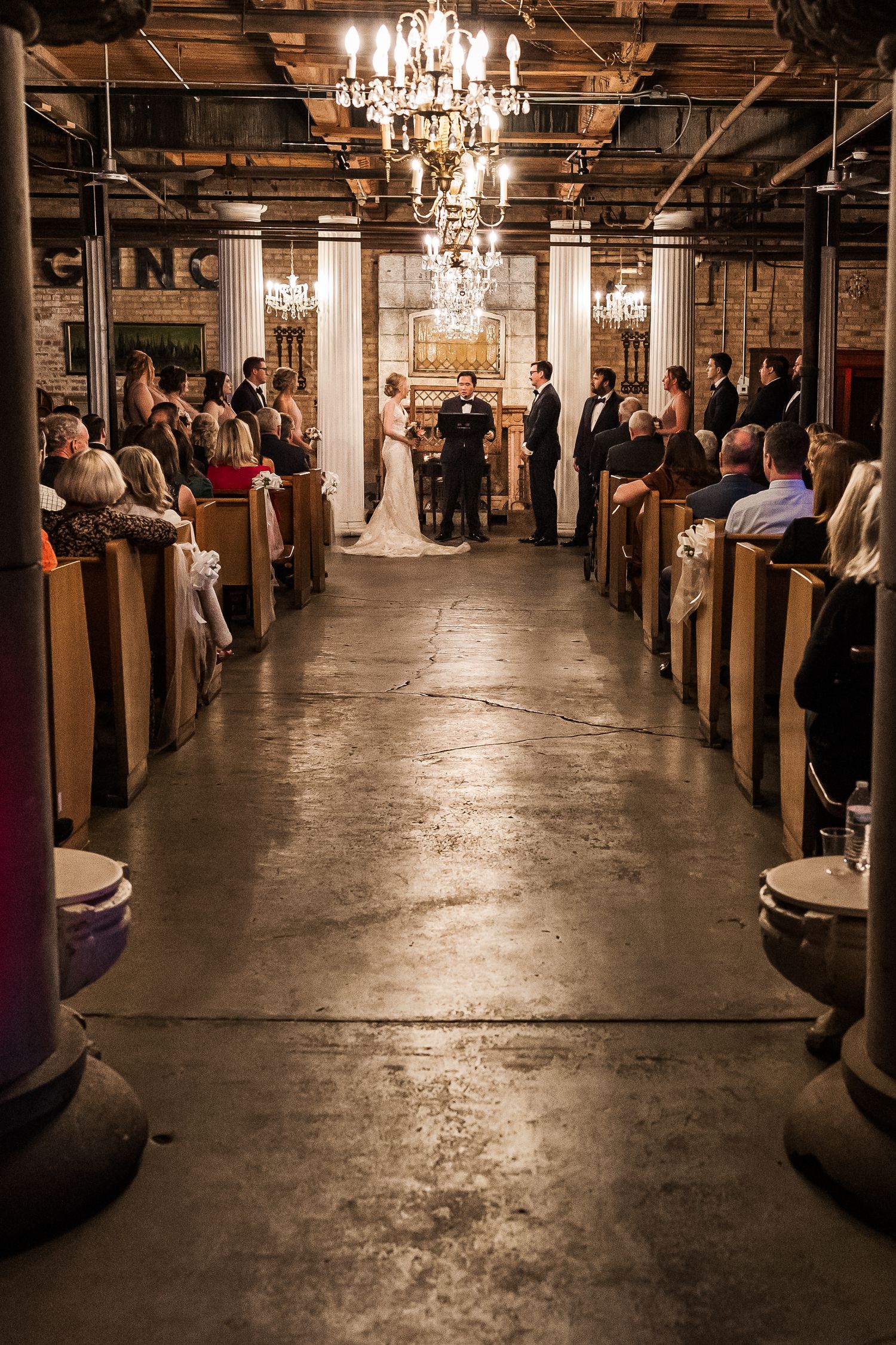 Wedding ceremony in an industrial venue with crystal chandeliers and wooden pews filled with guests.