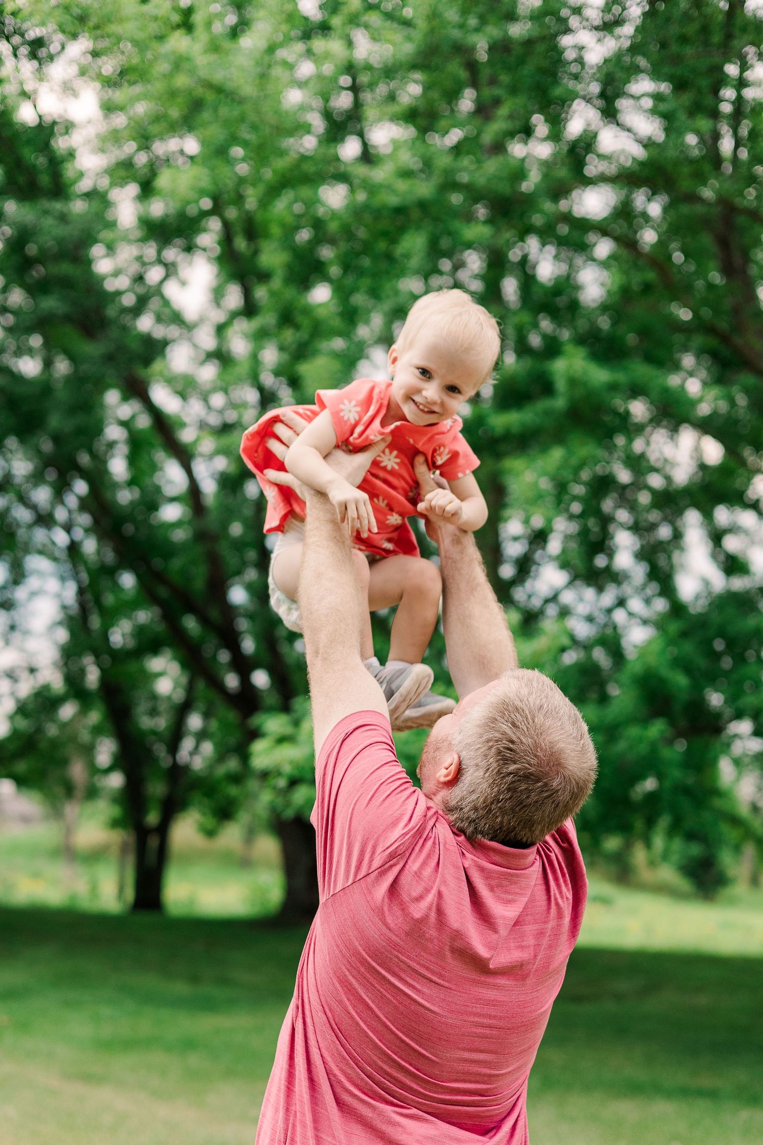 Parent lifting small baby high in the air against green trees background.