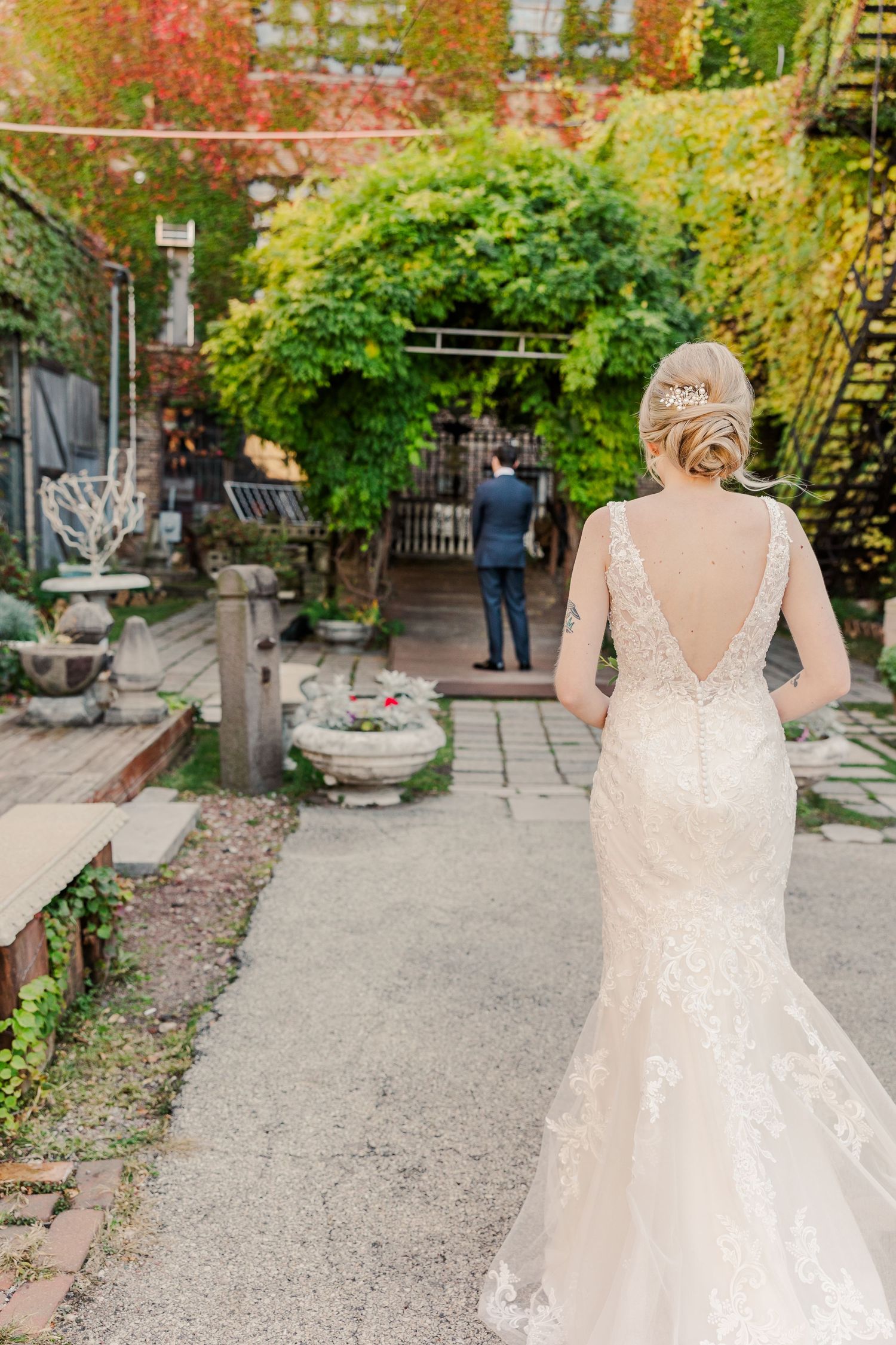Elegant bride in form-fitting lace dress walks down garden path with trailing train and deep v-back detail.