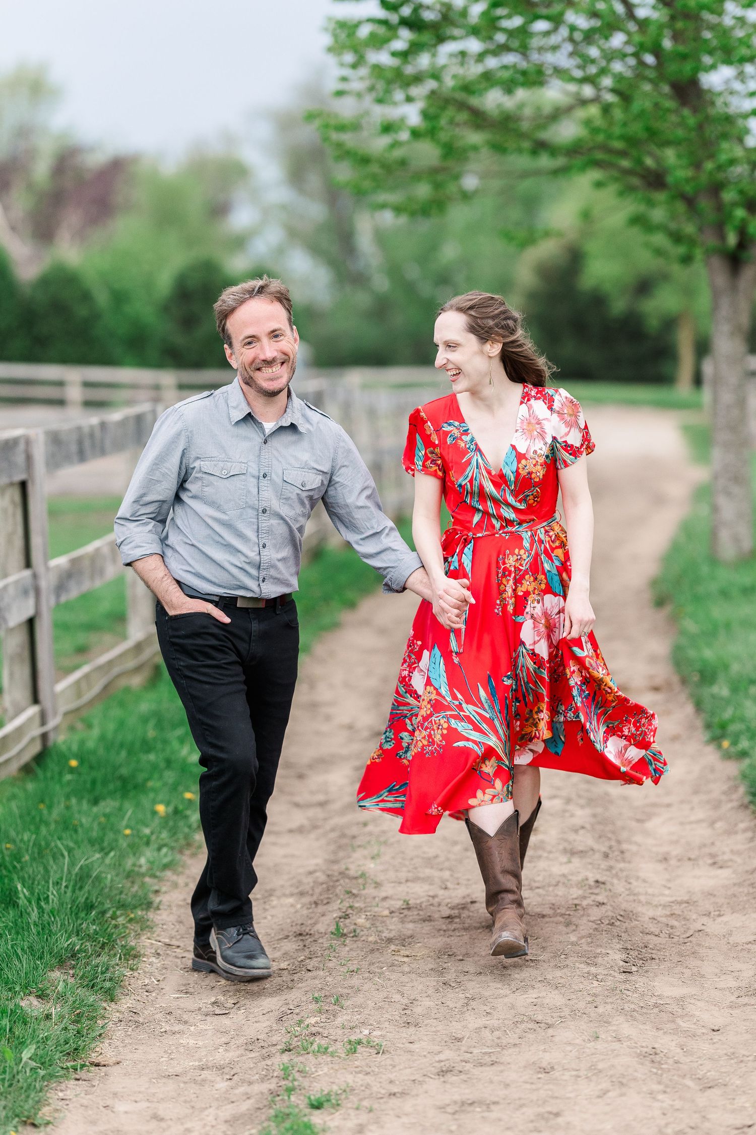 A couple walks hand in hand along a dirt path by wooden fences, with her red floral dress flowing.