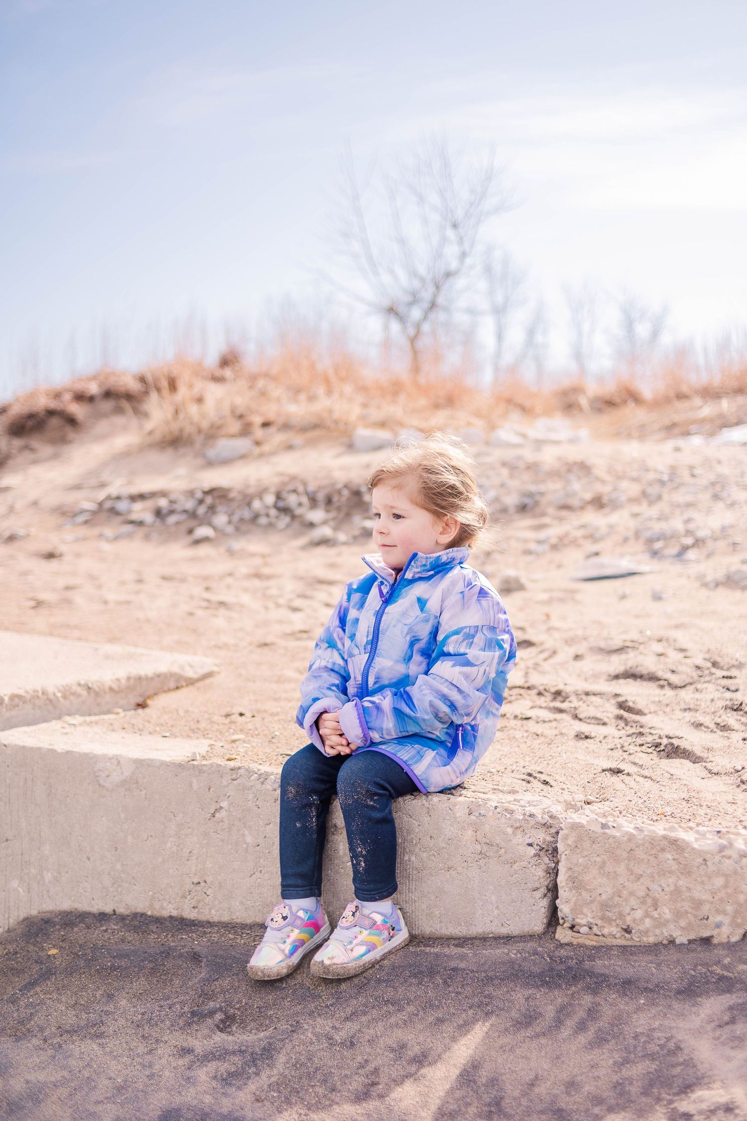 Someone wearing a tie-dye blue jacket sits on concrete blocks at a sandy beach.