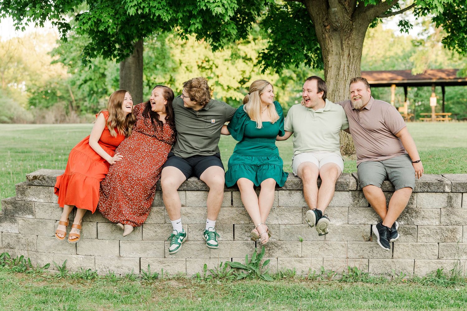 Friends sitting together on stone wall under tree at park during sunset.
