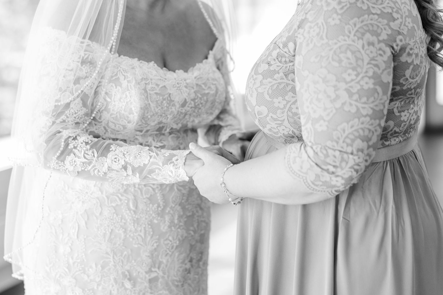 Black and white photo of a bride in a lace gown and a woman in a lace dress tenderly holding hands before the wedding.