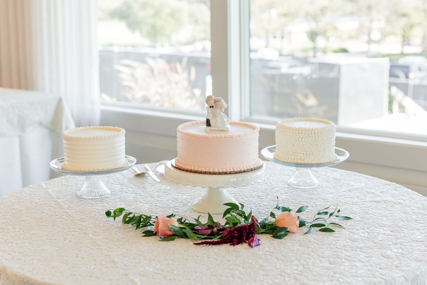 Wedding cake table in front of bright windows with a pink cake and cake topper, two white cakes, and a floral garland.