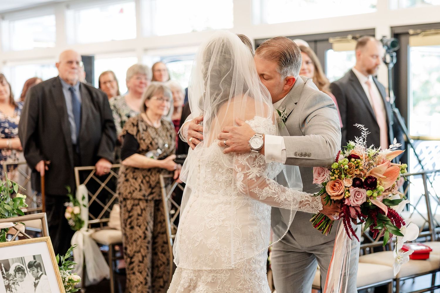 Bride in a lace gown and veil embraces a man in a gray suit during a wedding ceremony while guests look on and a bouquet rests nearby.