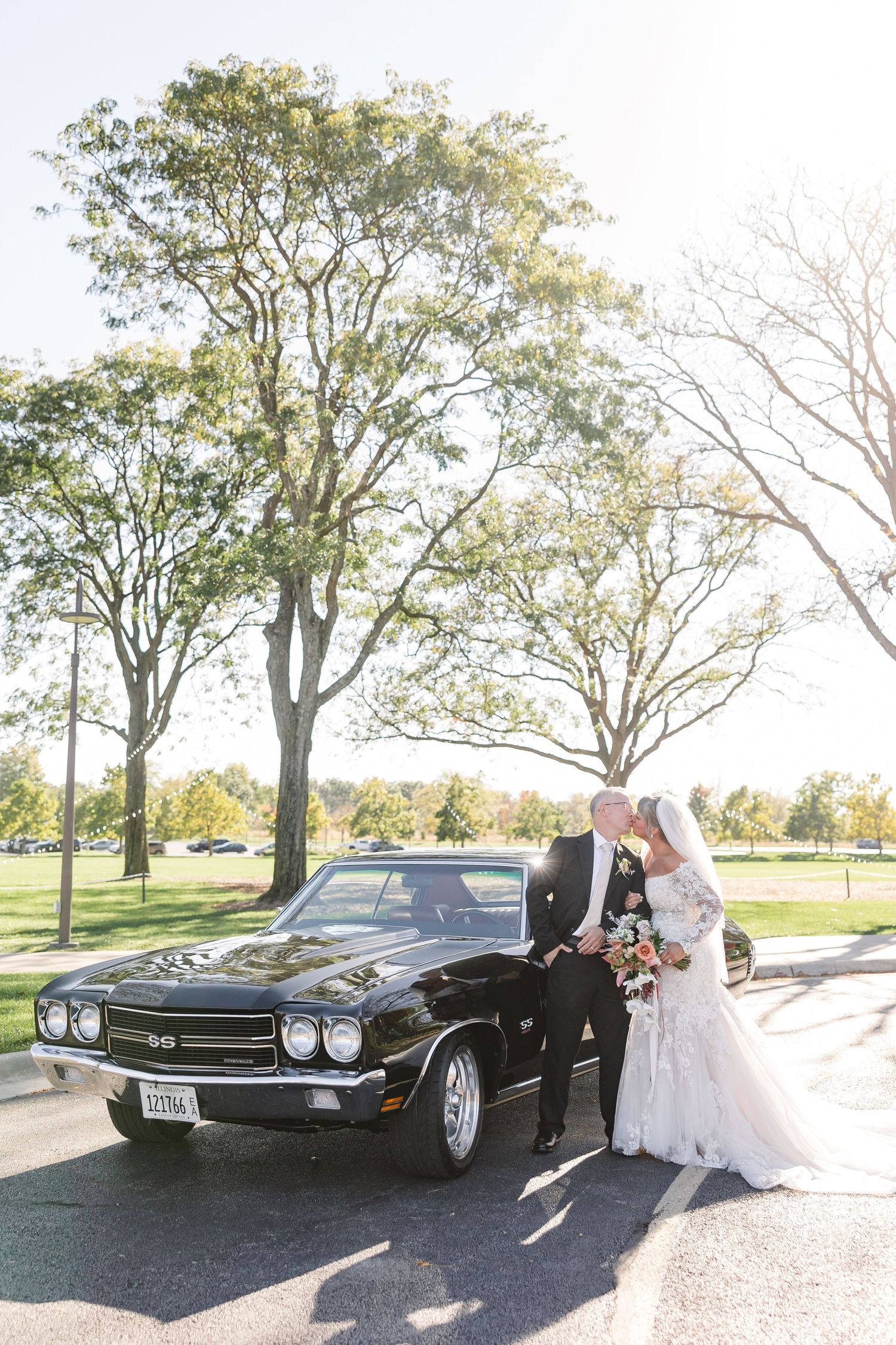 Bride in a lace gown and groom in a black suit kiss beside a classic black Chevelle muscle car under tall trees on a sunny day.