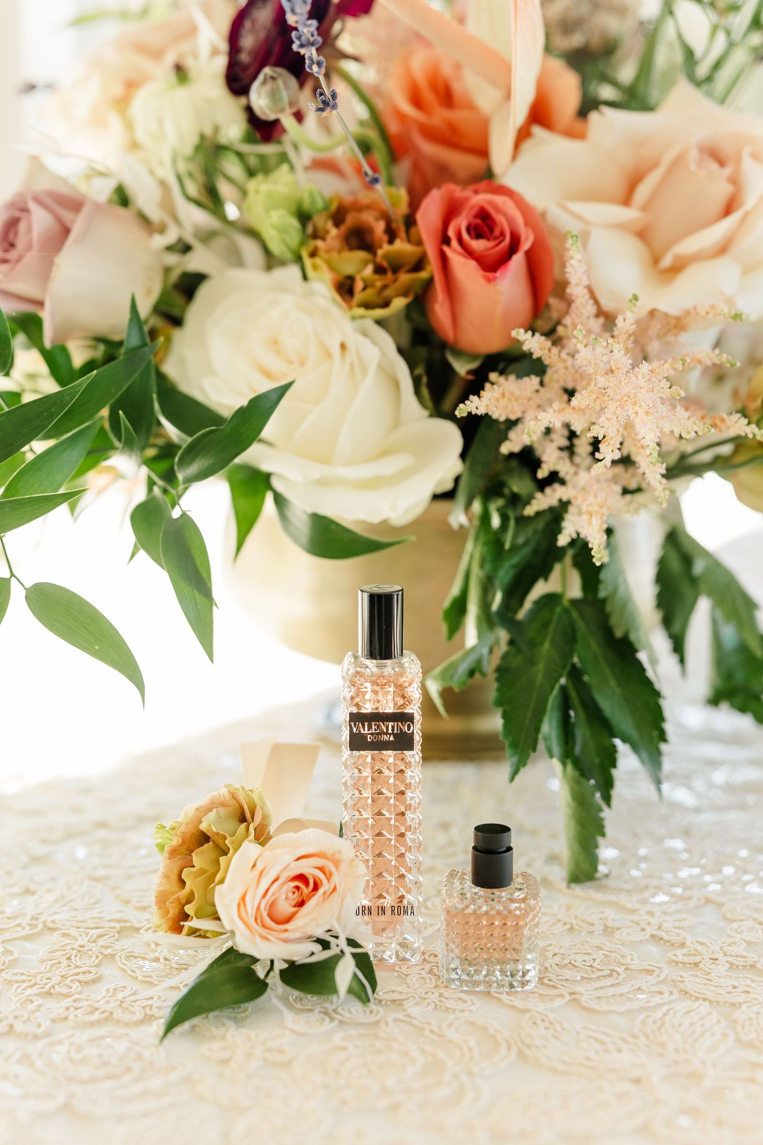 Valentino Donna perfume bottles beside a peach rose boutonniere and a bouquet of cream, blush and coral roses on a lace tablecloth.