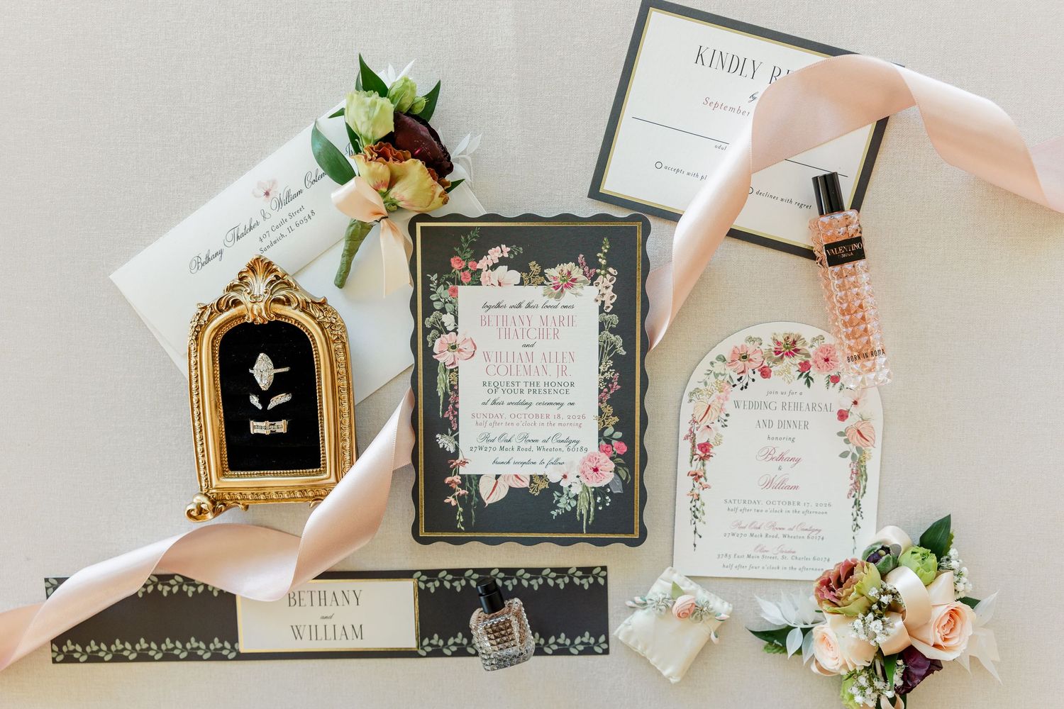Elegant floral wedding invitation suite with blush ribbon, gold frame, perfume bottle, and rose boutonniere styled on a white surface.