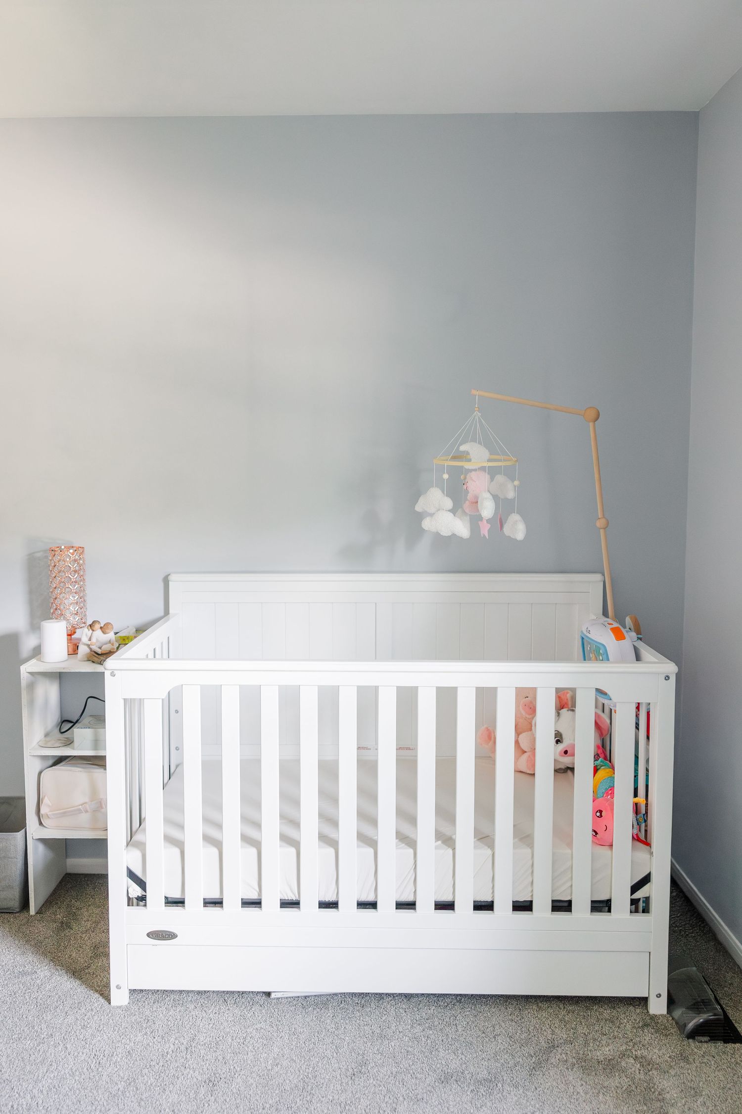 A white baby crib with a decorative mobile in a soft gray nursery room with carpet flooring.