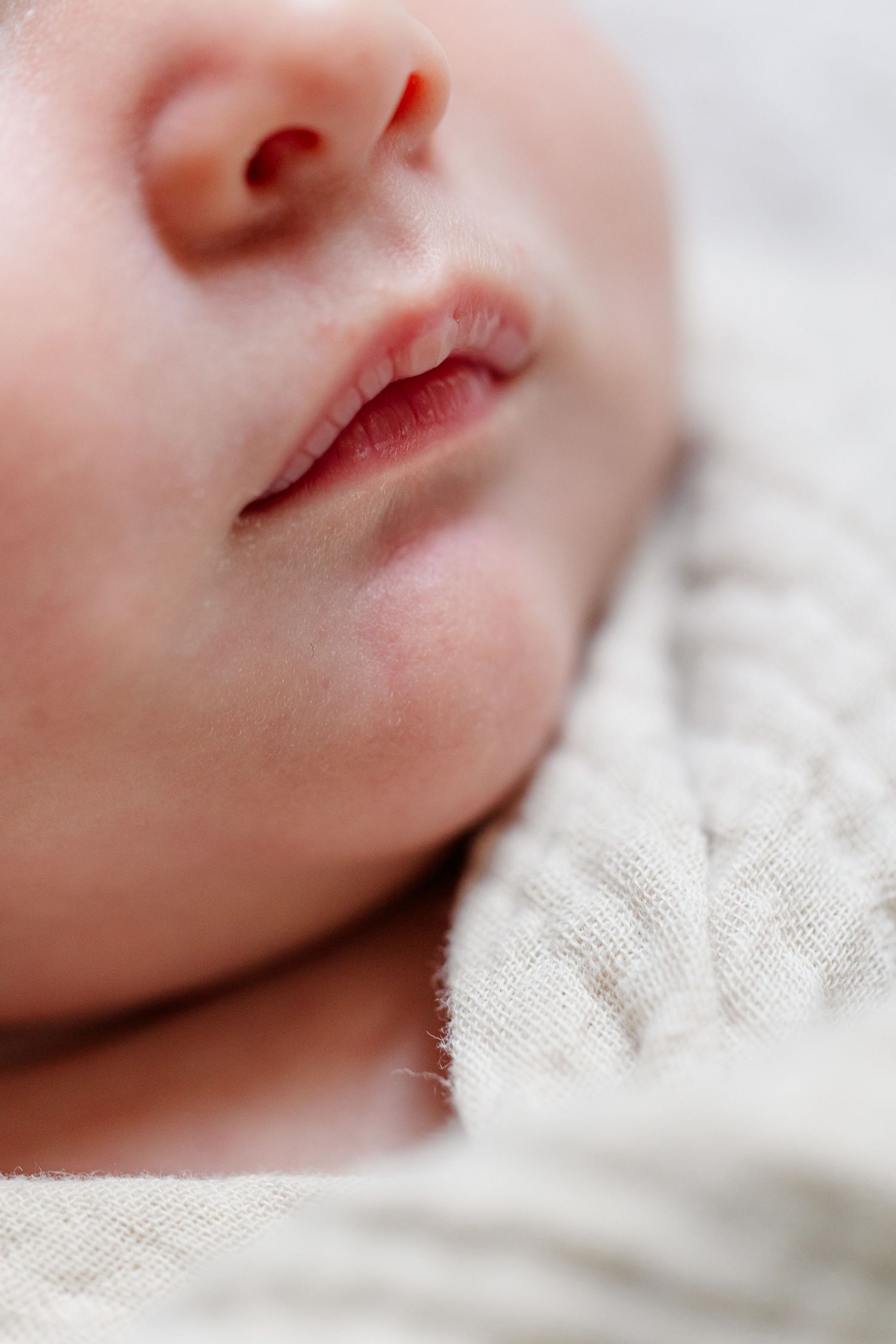Close-up of a peaceful sleeping newborn baby's face, showing tiny lips and nose, wrapped in a soft cream knit blanket.