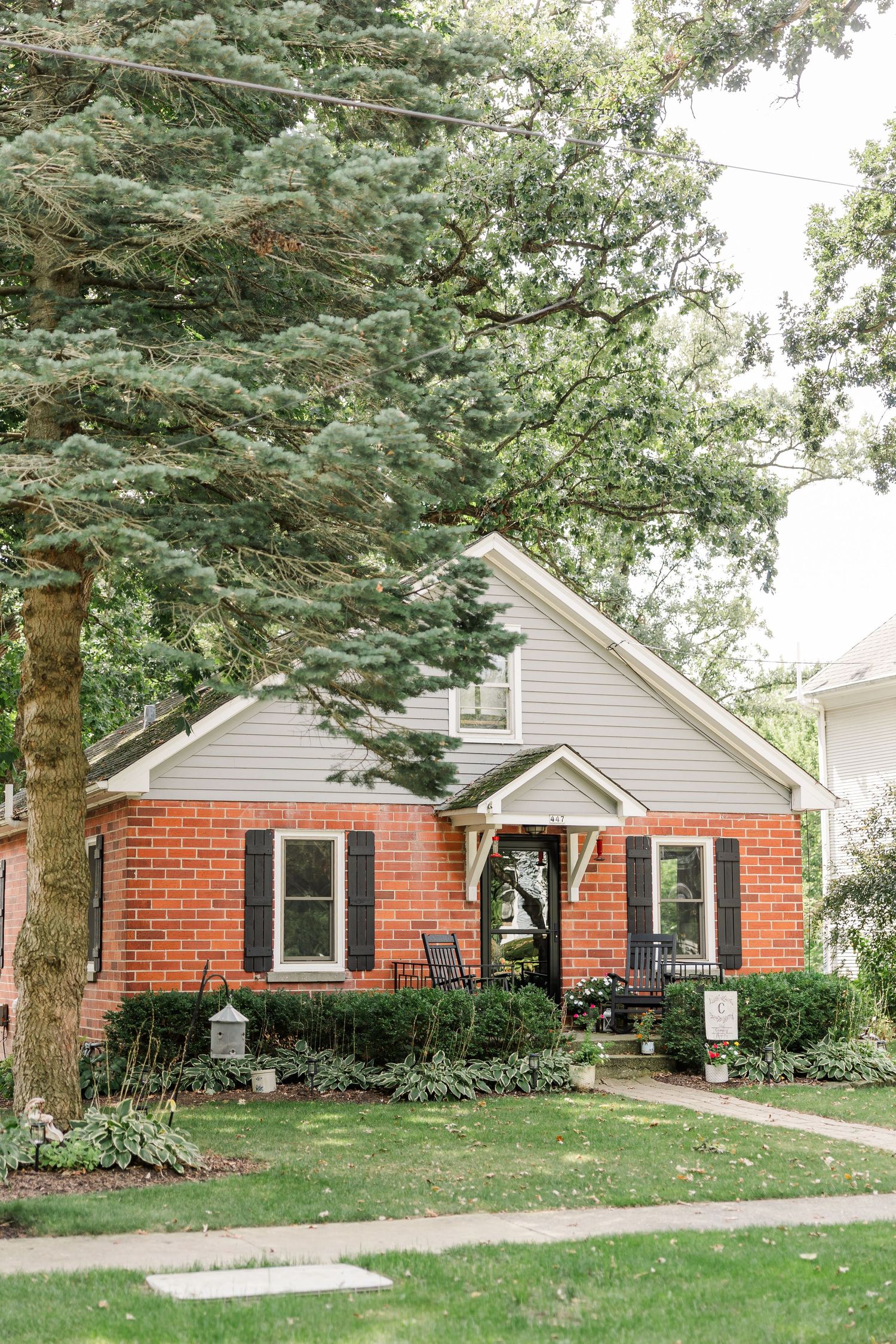 Charming red brick cottage home with black shutters surrounded by lush mature trees and manicured landscaping.