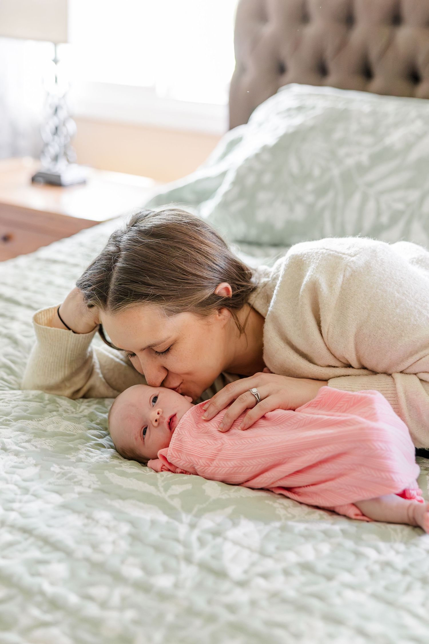 A mother tenderly kisses her swaddled newborn baby in pink while lying on a bed.