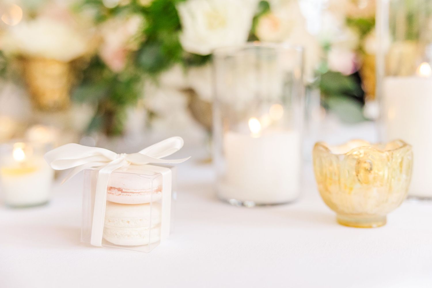 Favor boxes and gold votive holders on white tablecloth with soft floral decorations in background.