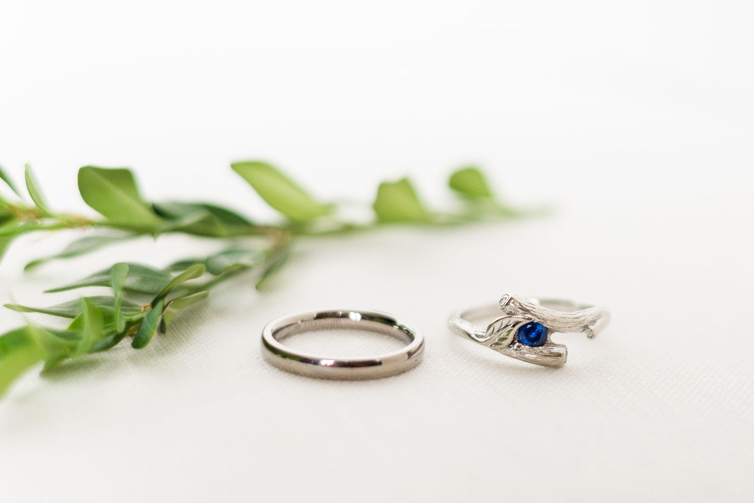 Wedding rings and engagement ring with blue stone displayed with green foliage.