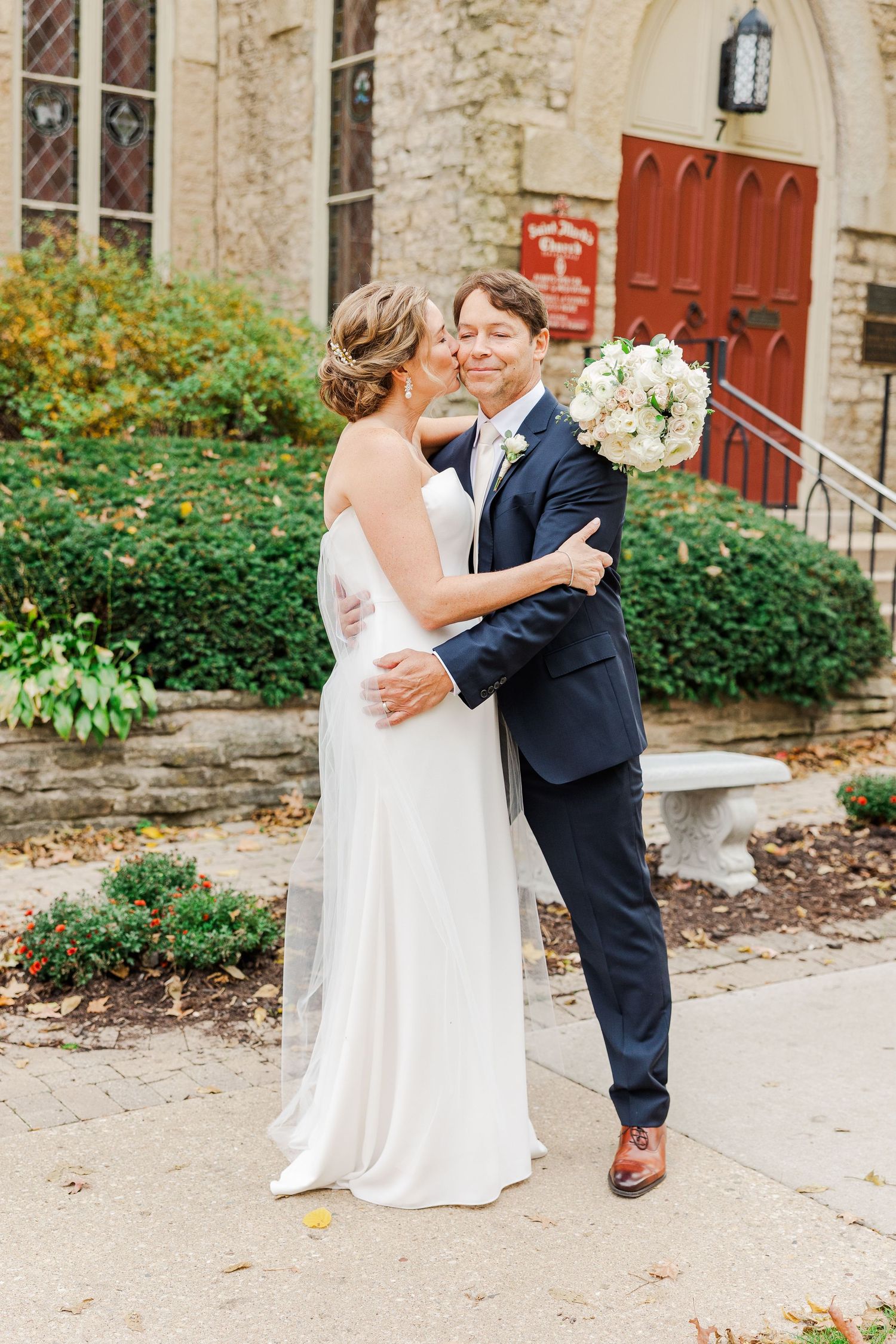 Wedding couple embracing outside stone church with red door in autumn.