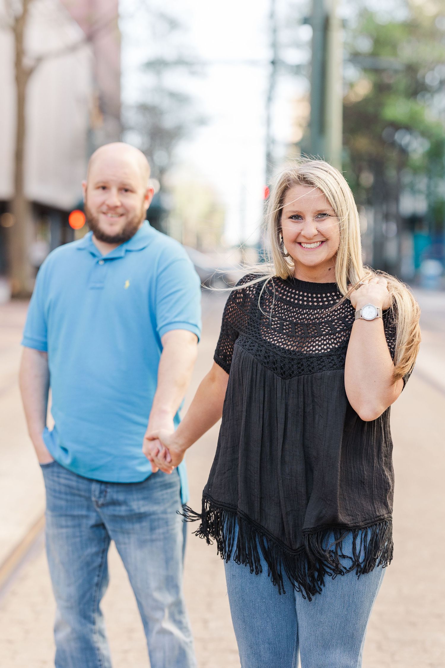 A couple poses casually on an urban street with vintage lampposts and trees.