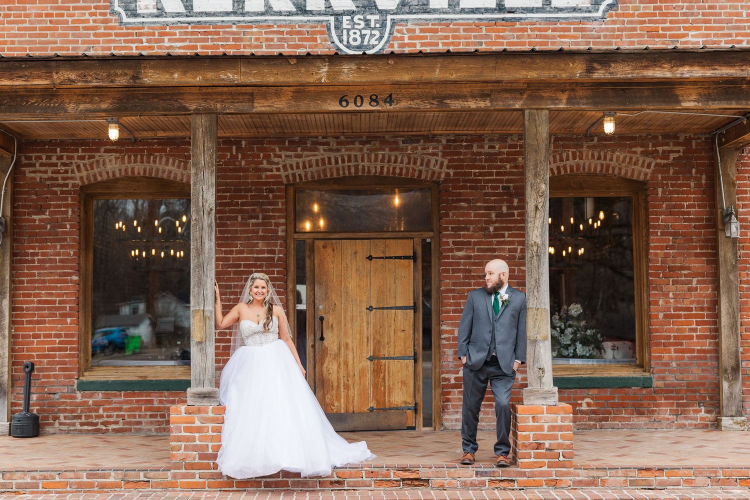 Wedding couple poses in front of rustic brick storefront with wooden door.