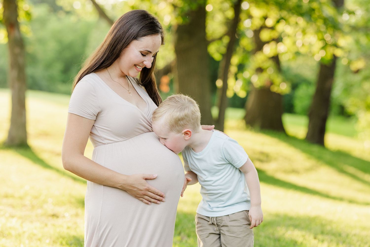 A pregnant woman tenderly holds her baby bump while standing in a sunlit forest with soft glowing bokeh effect.