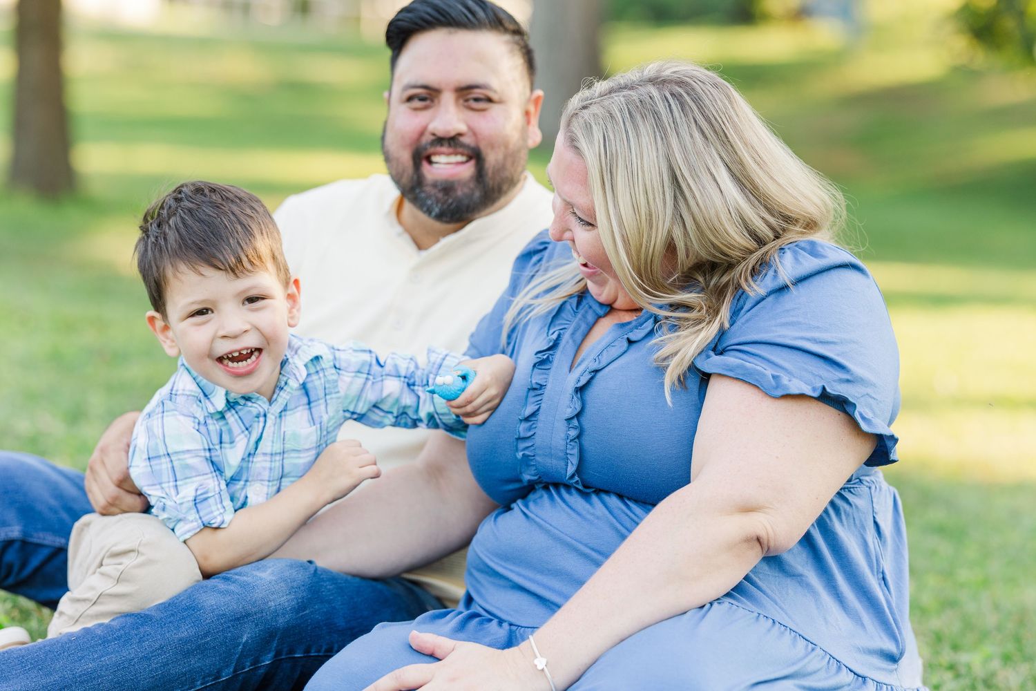 A family sits together and laughs on green grass during a sunny outdoor portrait session.Sorg family portraits at Shamrock Hills Park in Cry