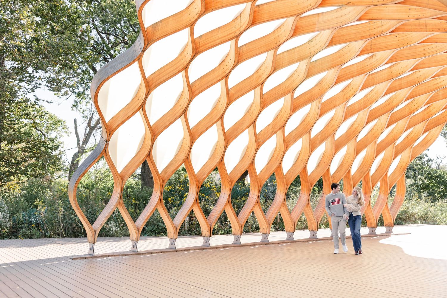 Stunning wooden lattice pavilion with organic honeycomb design on a deck, surrounded by lush green trees.