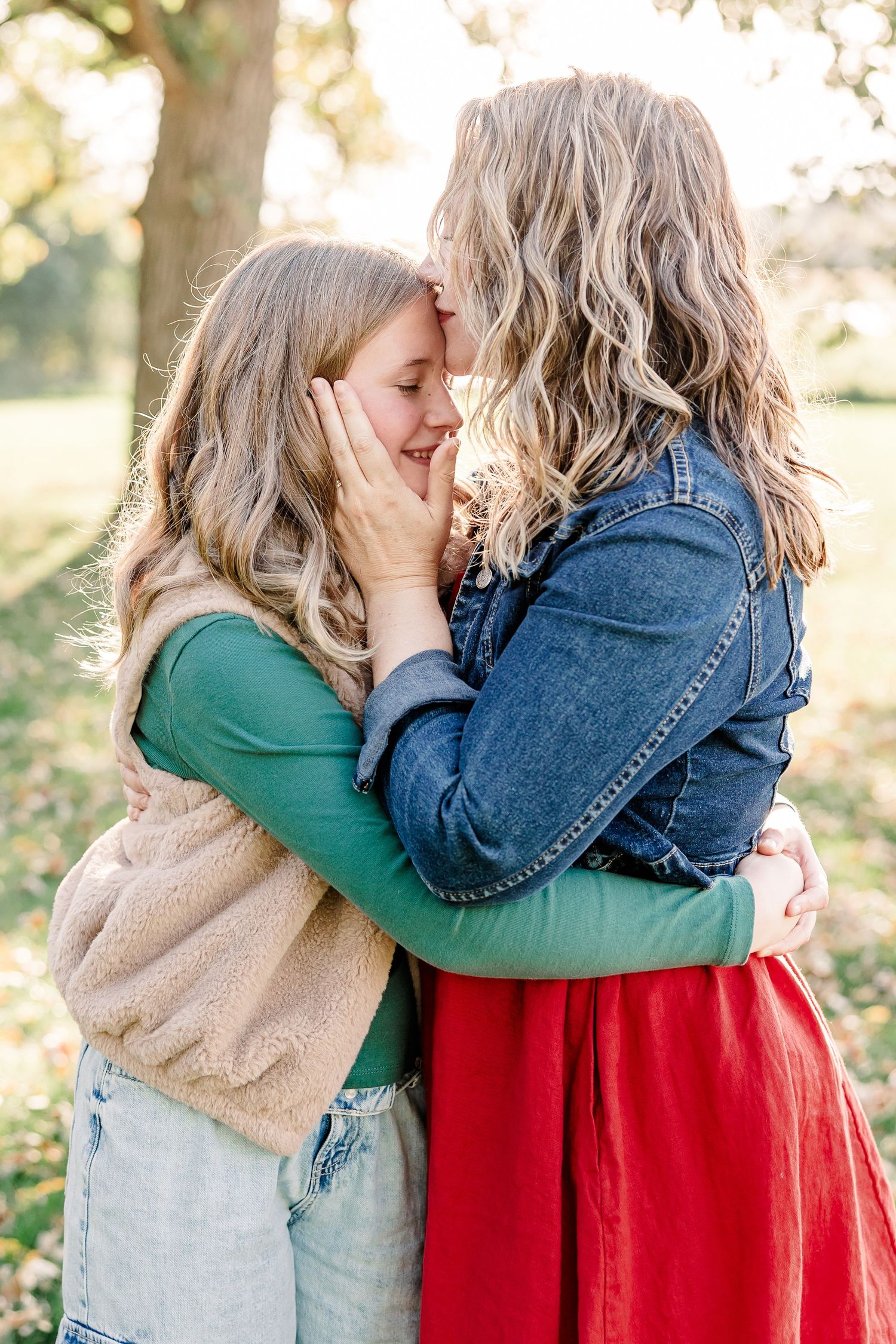 Two women embrace outdoors in soft golden light as one gently kisses the other's forehead, wearing a denim jacket and a red skirt.