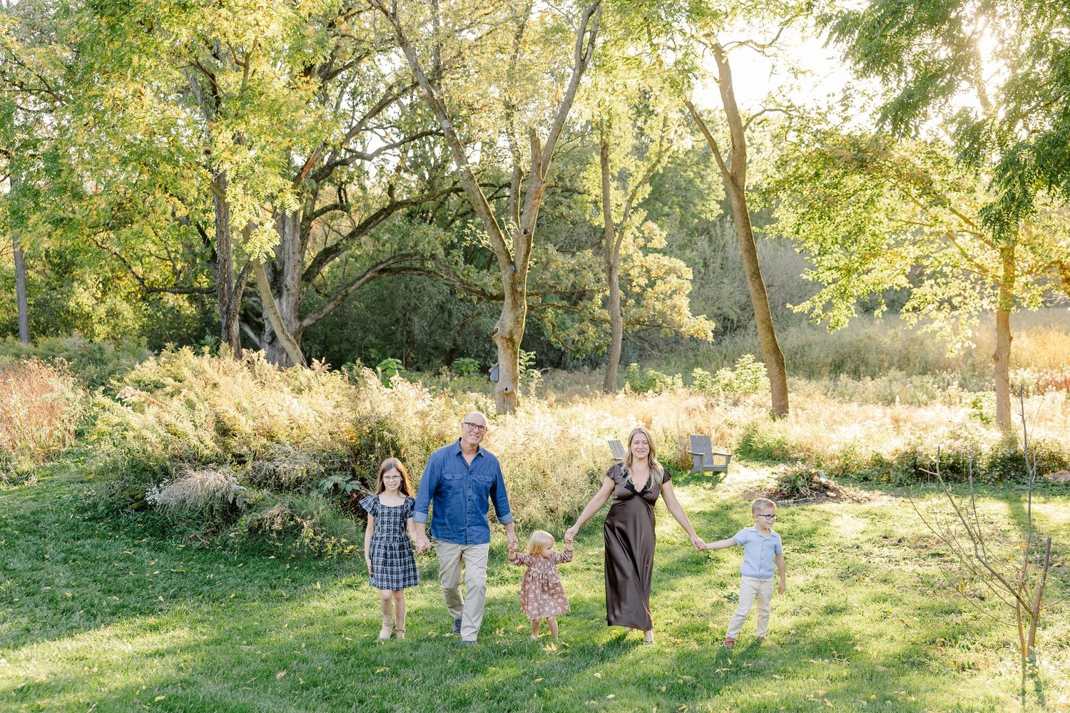 Family of five walking hand in hand beneath sunlit trees in their St. Charles, IL backyard.