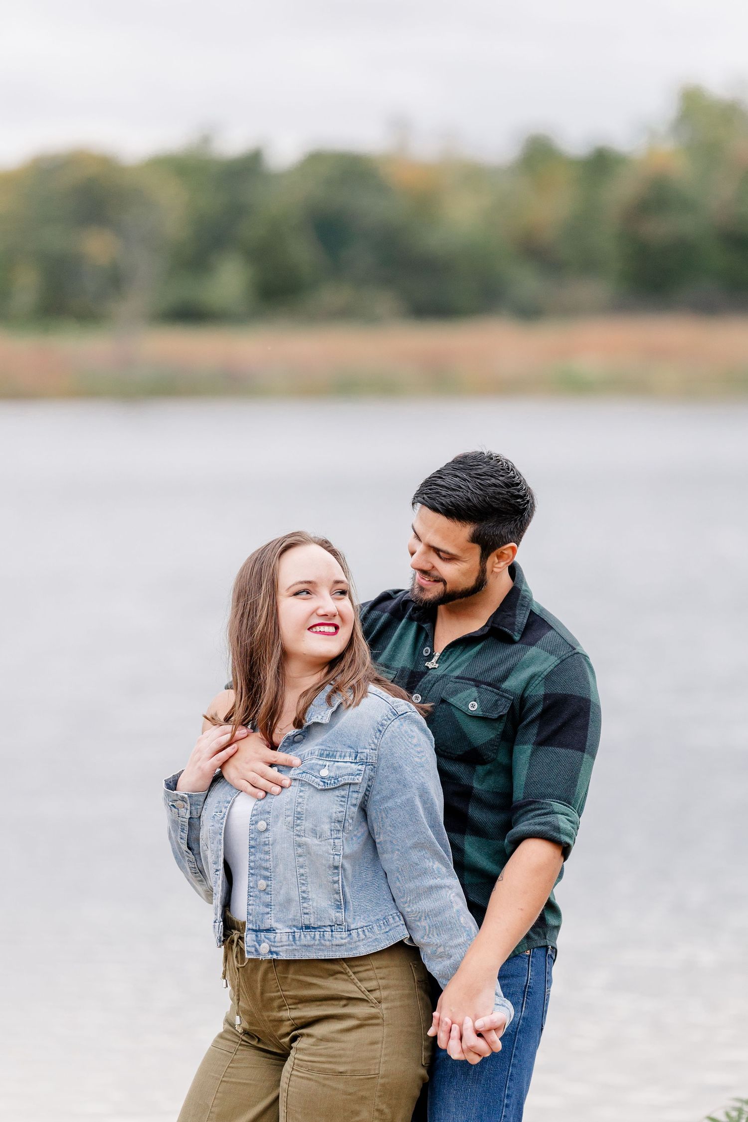 Smiling couple embracing by a calm lake, the woman in a denim jacket and the man in a green plaid flannel shirt holding her.