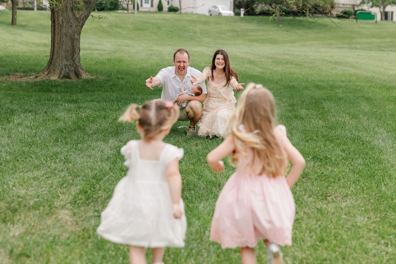Two little girls in pink dresses run toward their smiling parents sitting on a lush green lawn.