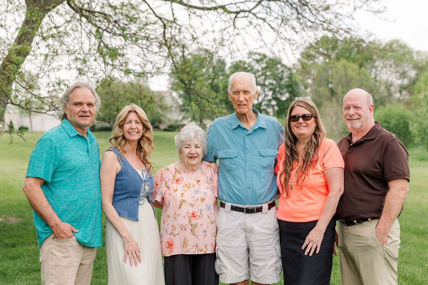 Six family members smiling together outdoors on a green lawn beneath leafy trees during a warm spring day.