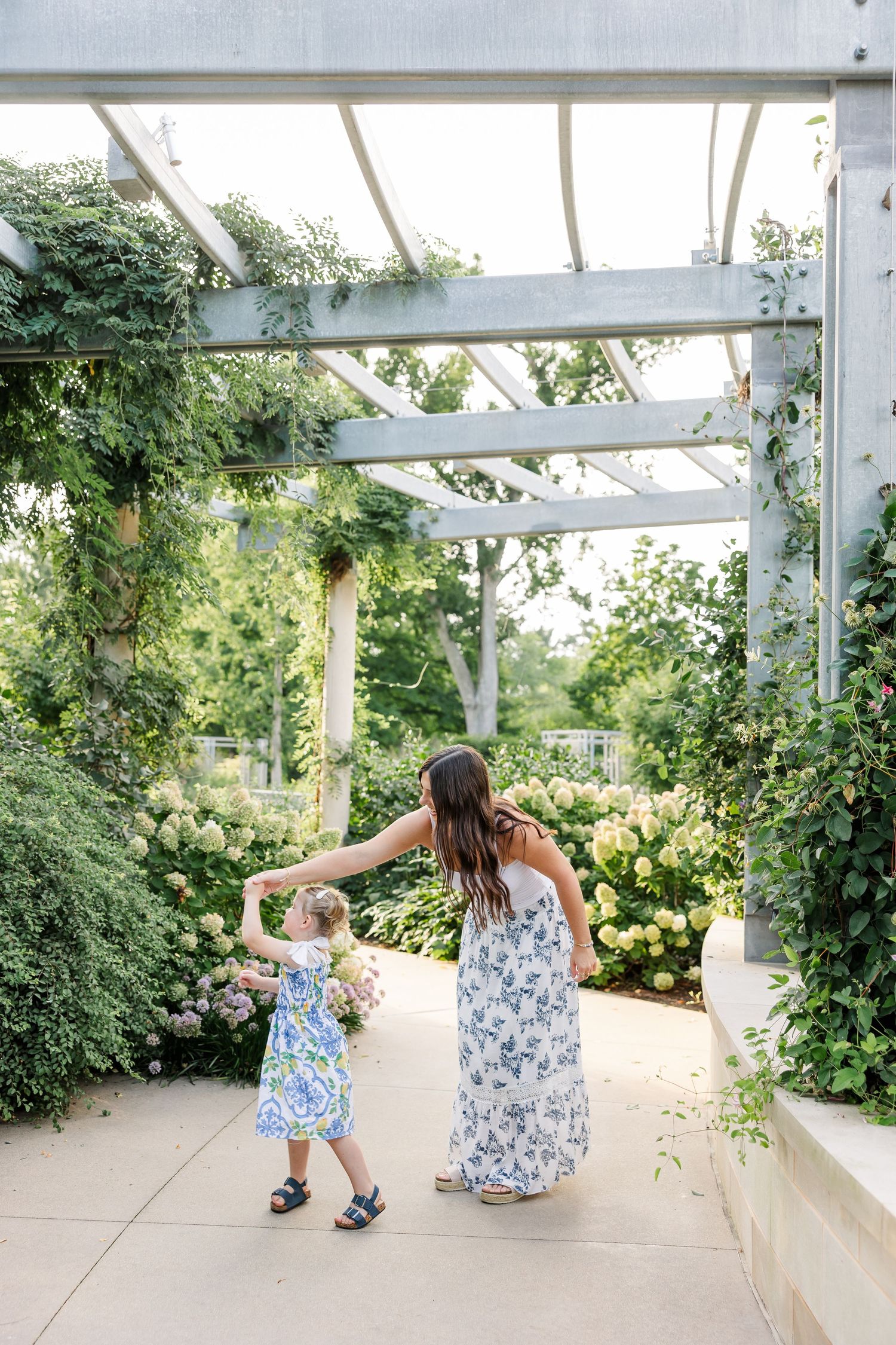 Woman in floral dress holds hands with toddler girl under garden pergola with roses.