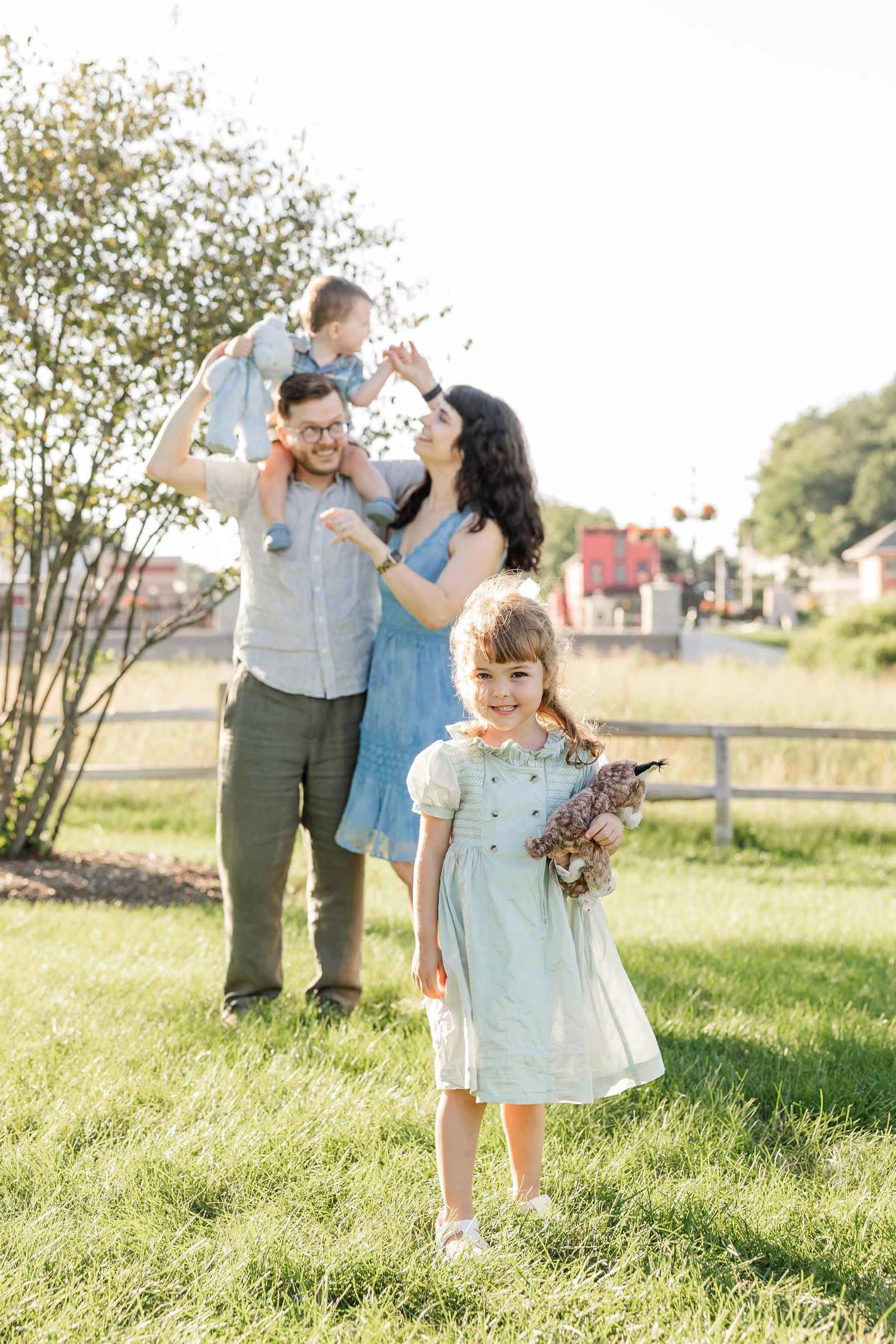A joyful family of four poses outdoors on green grass, with dad lifting baby while mom and young daughter smile in blue dresses.