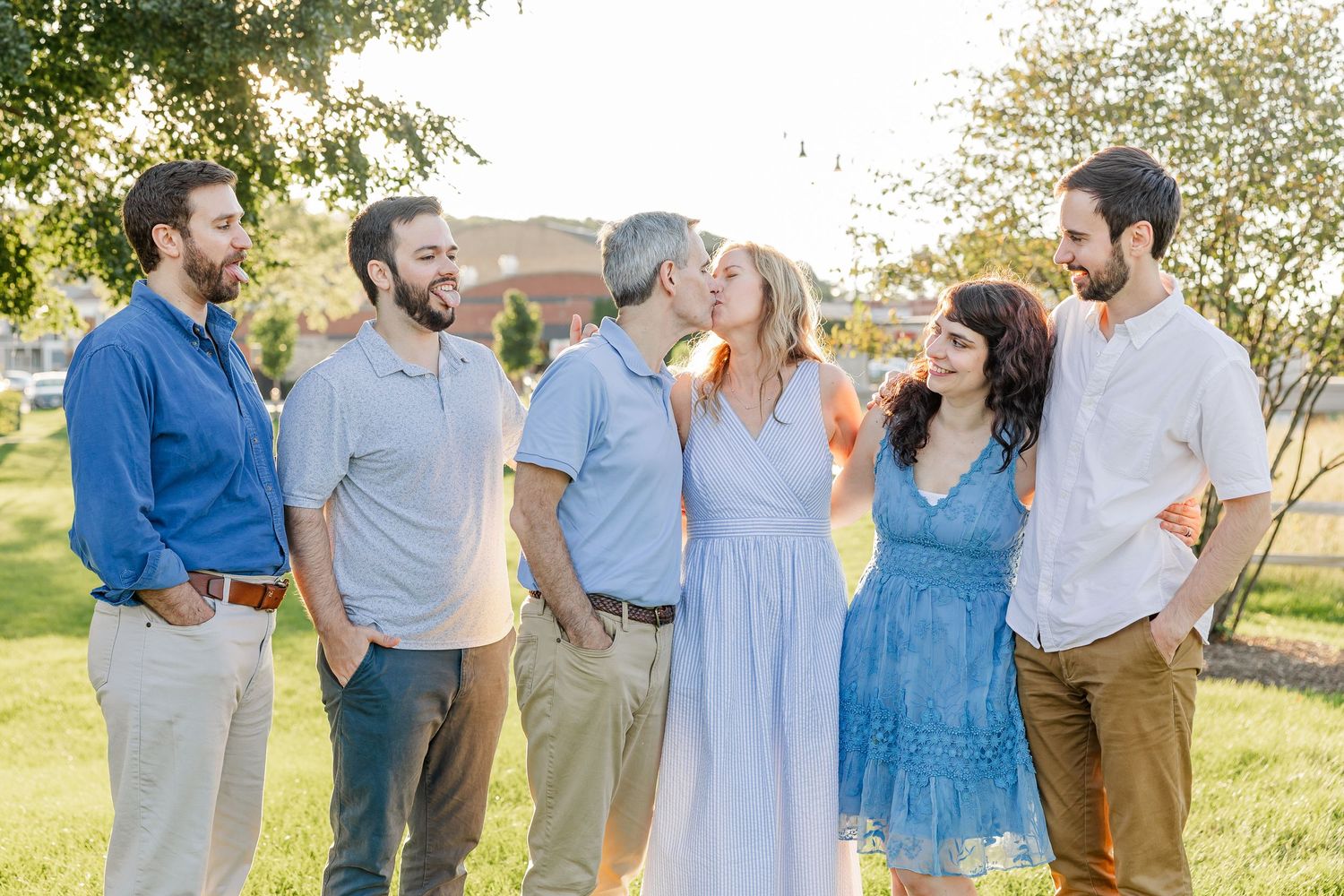 Family group of six adults standing outdoors in golden light, with couple sharing a kiss in center.
