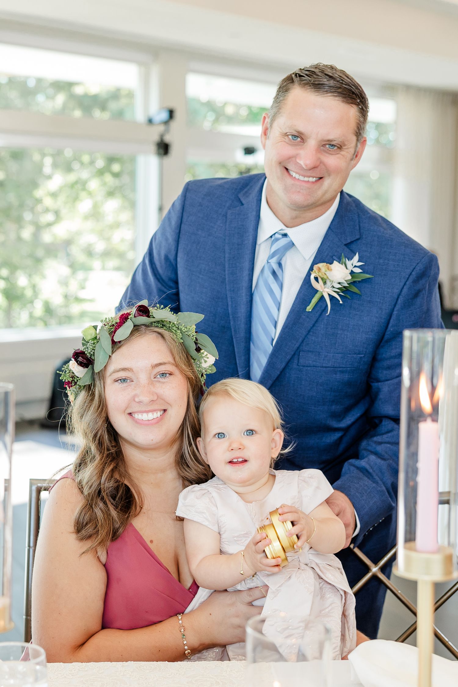 Smiling man in a blue suit with a boutonniere stands behind a woman in a floral crown holding a baby at a bright wedding reception table.