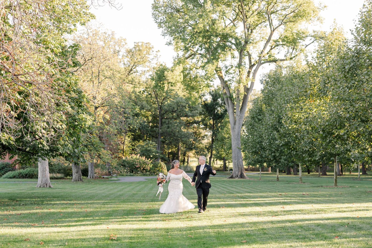 Bride in a white gown and groom in a dark suit walk hand in hand across a sunlit green lawn surrounded by tall trees at an outdoor wedding.