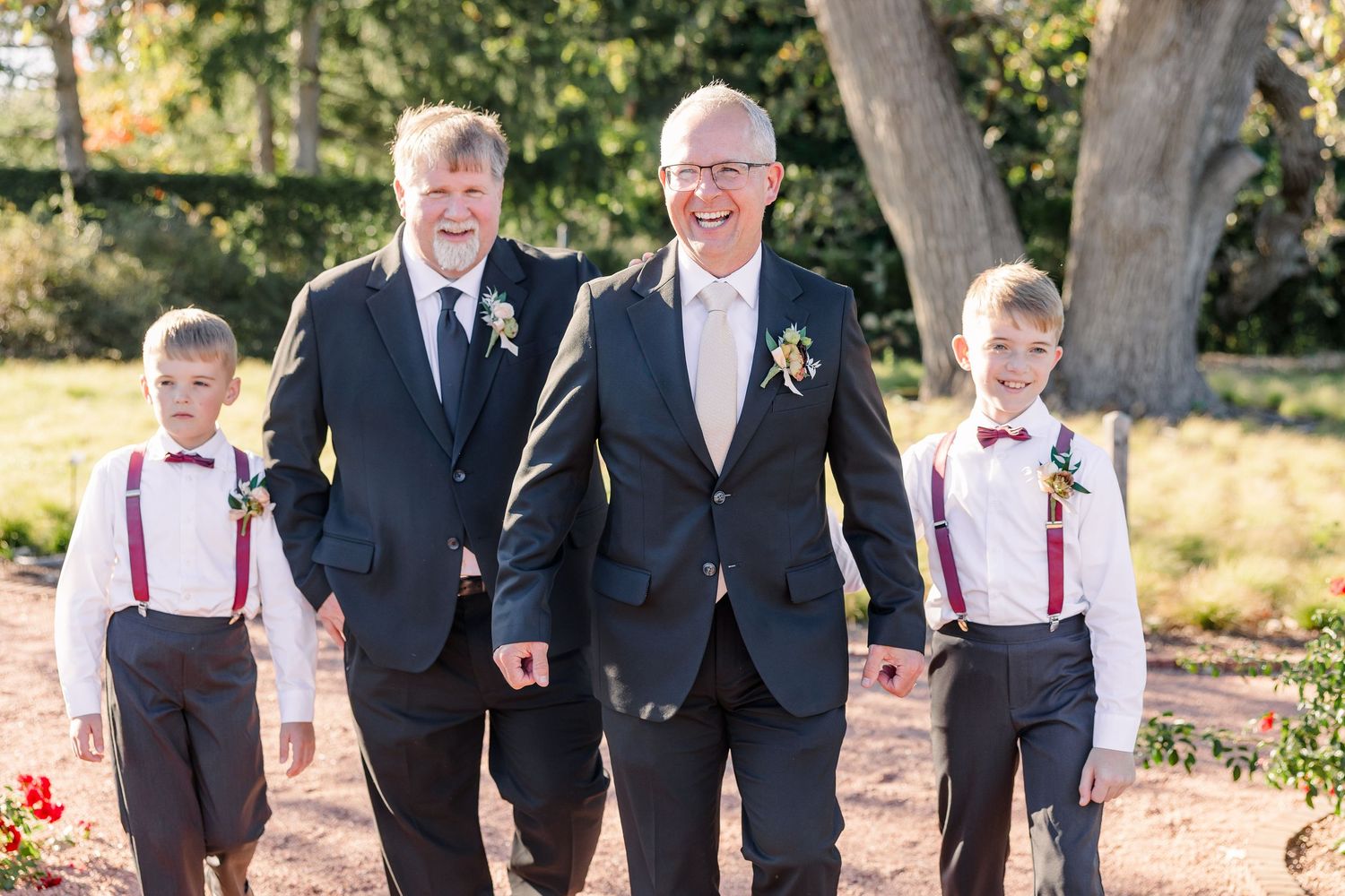 Two men in dark suits with boutonnieres walk with two boys in suspenders and bow ties along a garden path at a wedding.
