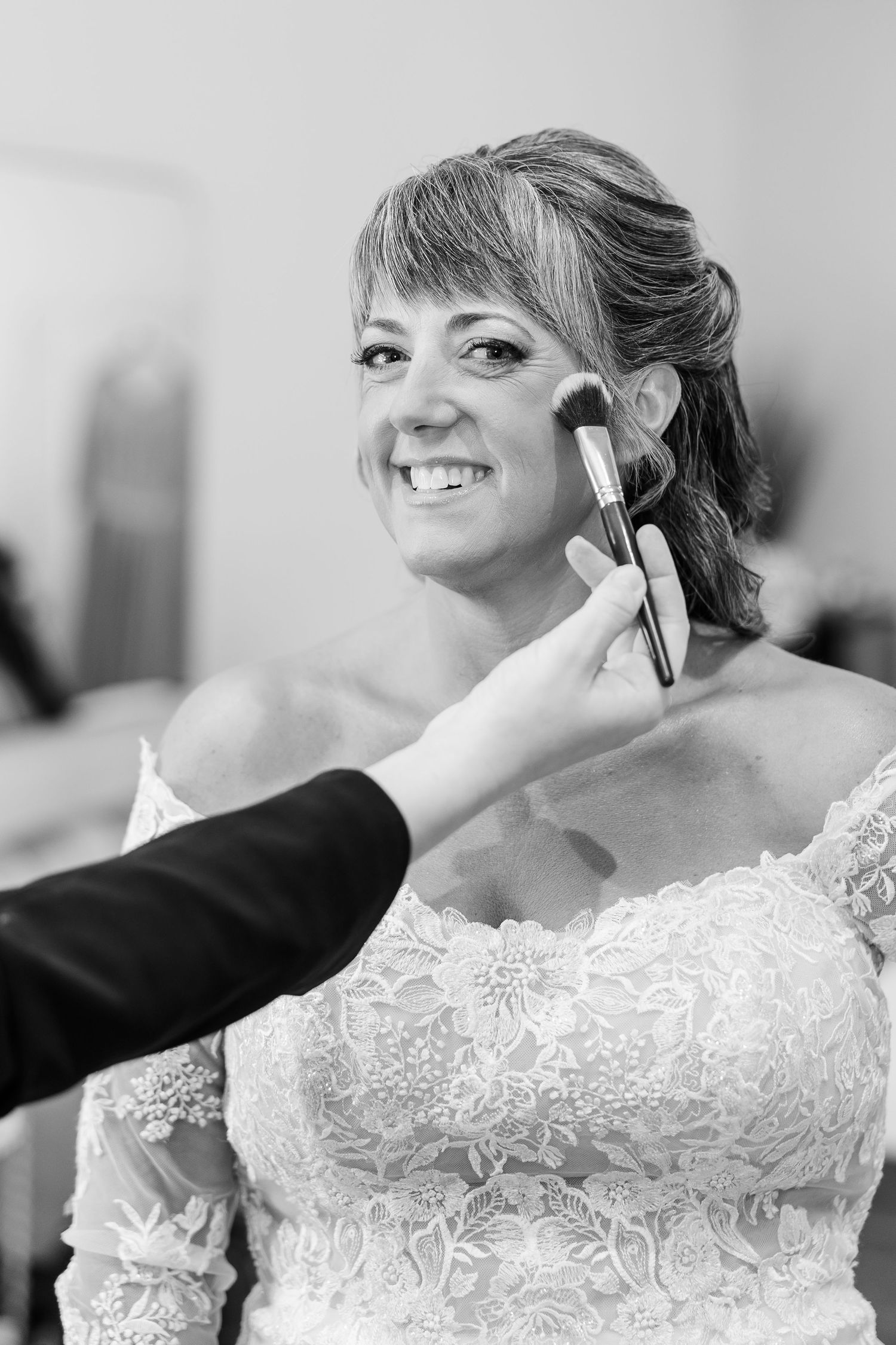 Black and white photo of a smiling bride in a lace off-shoulder gown having blush applied with a makeup brush.