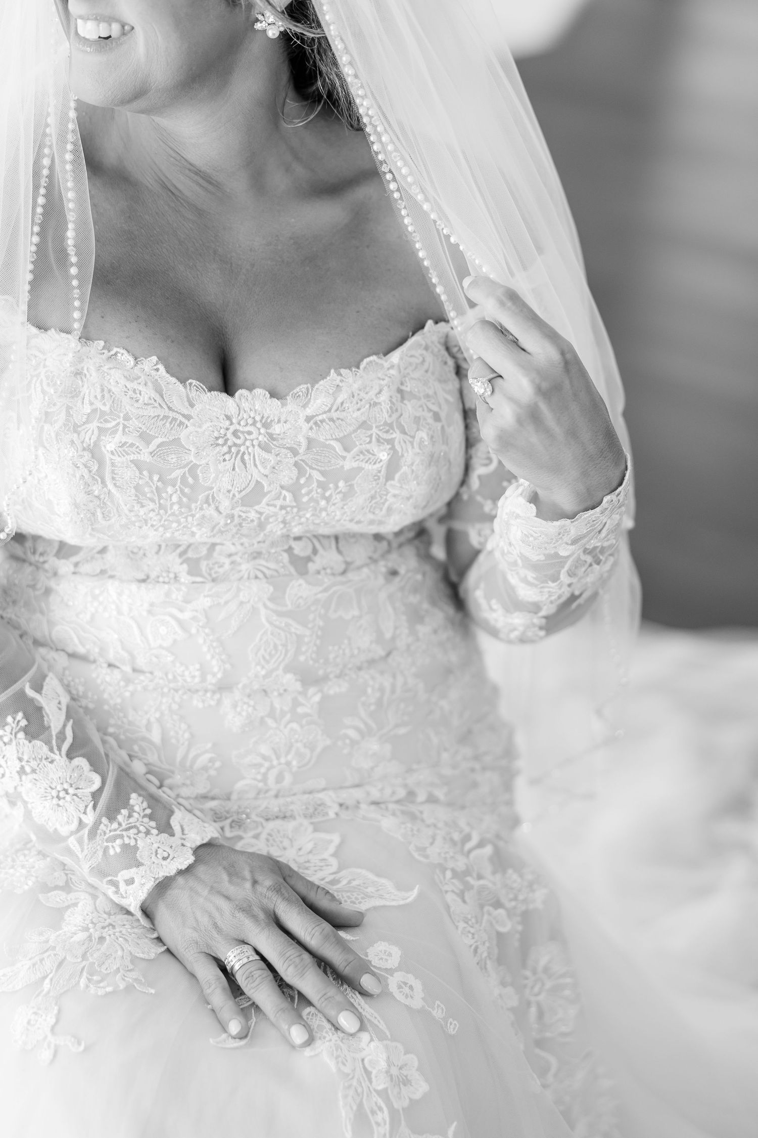 Black and white photo of a bride in an off-the-shoulder lace wedding gown and pearl-trimmed veil, gently holding her veil.