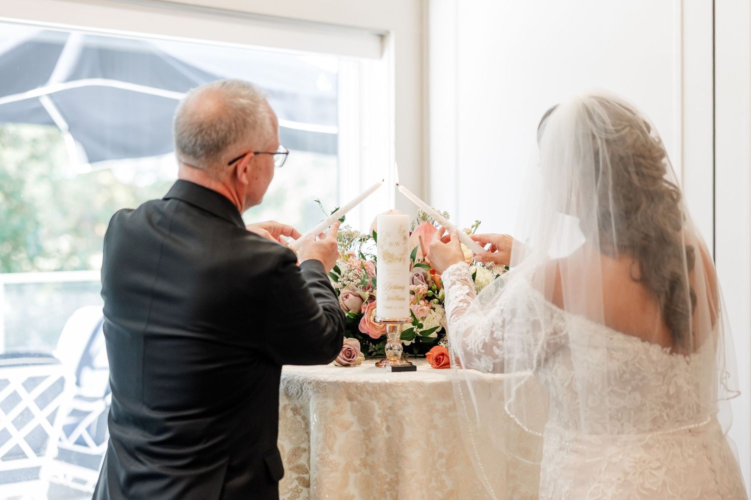 A bride in a lace gown and veil and an officiant in a black suit stand at a table during a wedding unity candle ceremony.