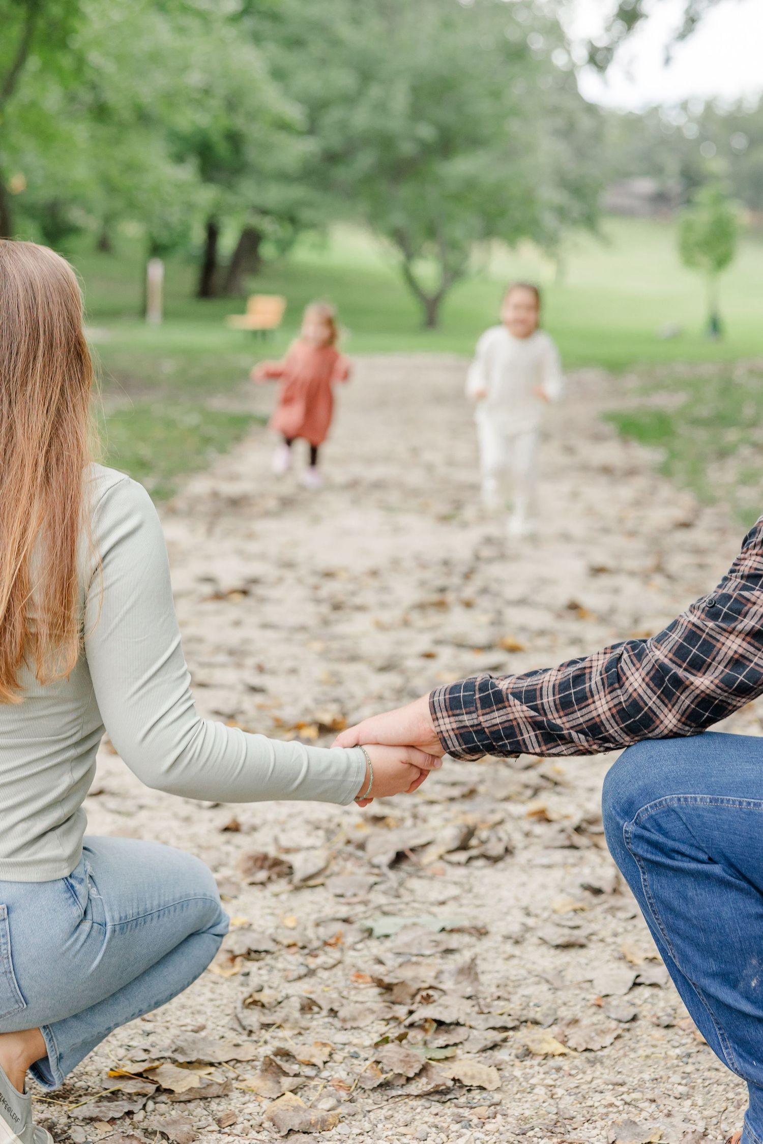 A couple holds hands while crouching as two children run toward them on a leaf-covered path in a green park.