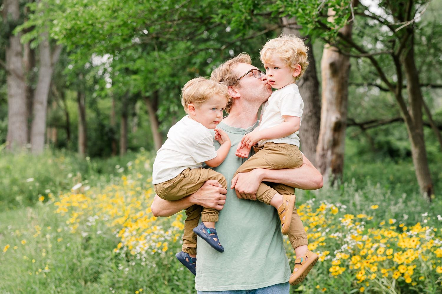 Parent holding two young children in a sunny wildflower meadow surrounded by lush green trees.