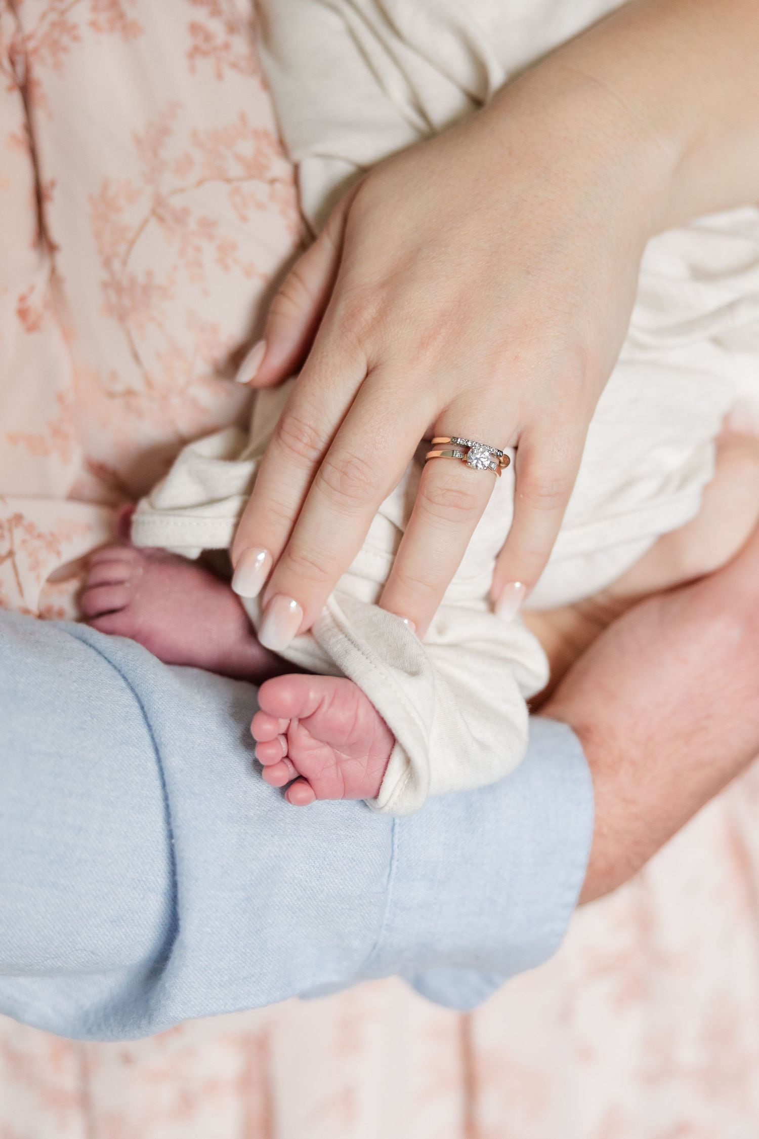 Close-up of mother's hand with diamond ring gently holding newborn baby's tiny feet in white swaddle.
