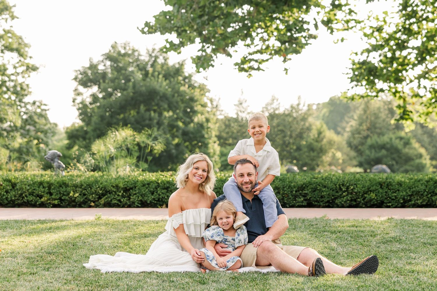 Happy family of four sitting on grass in a sunny park, parents smiling with two young children.